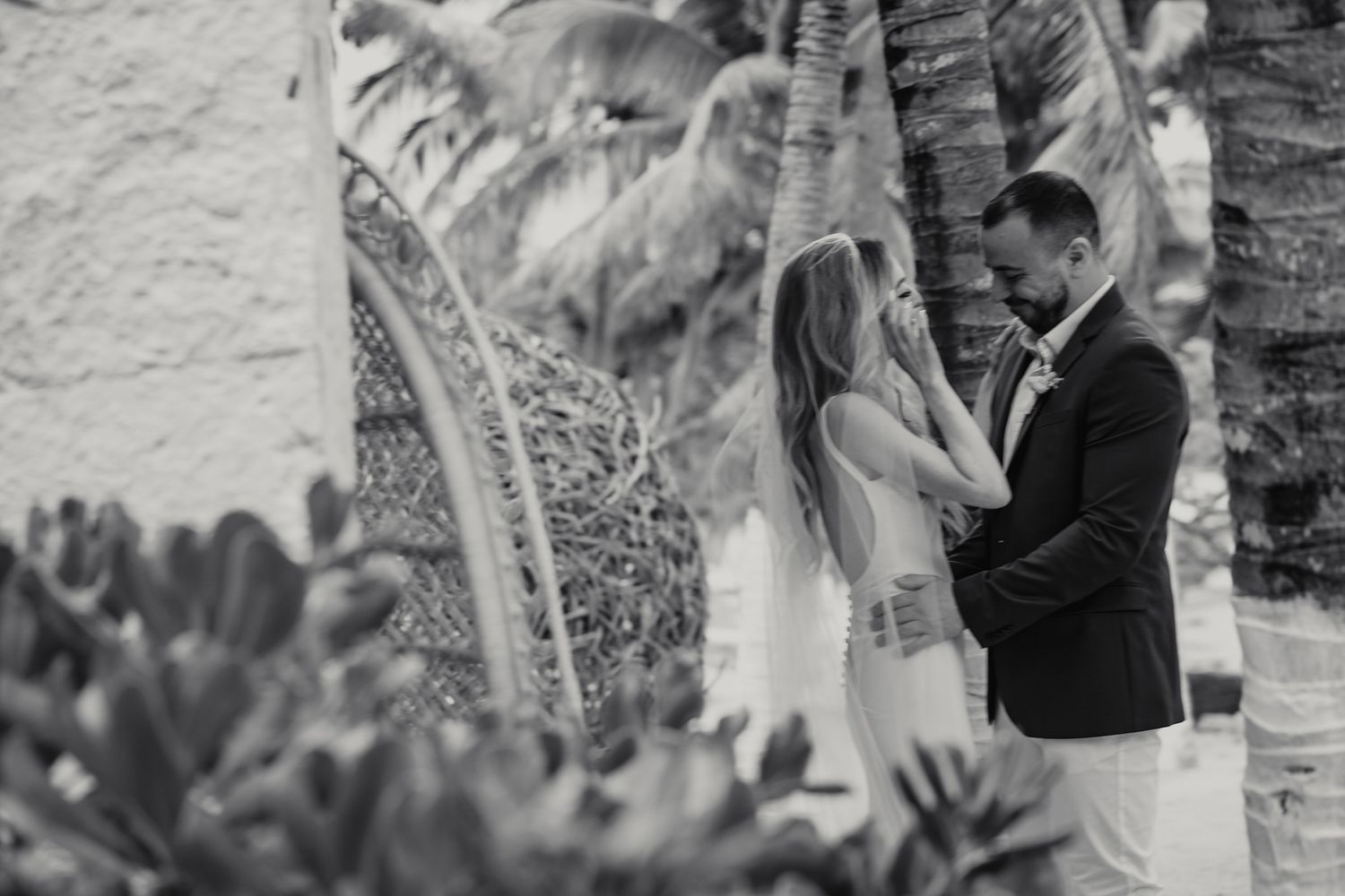 Black and white photo of bride and groom holding hands near tropical palm trees at beach wedding.