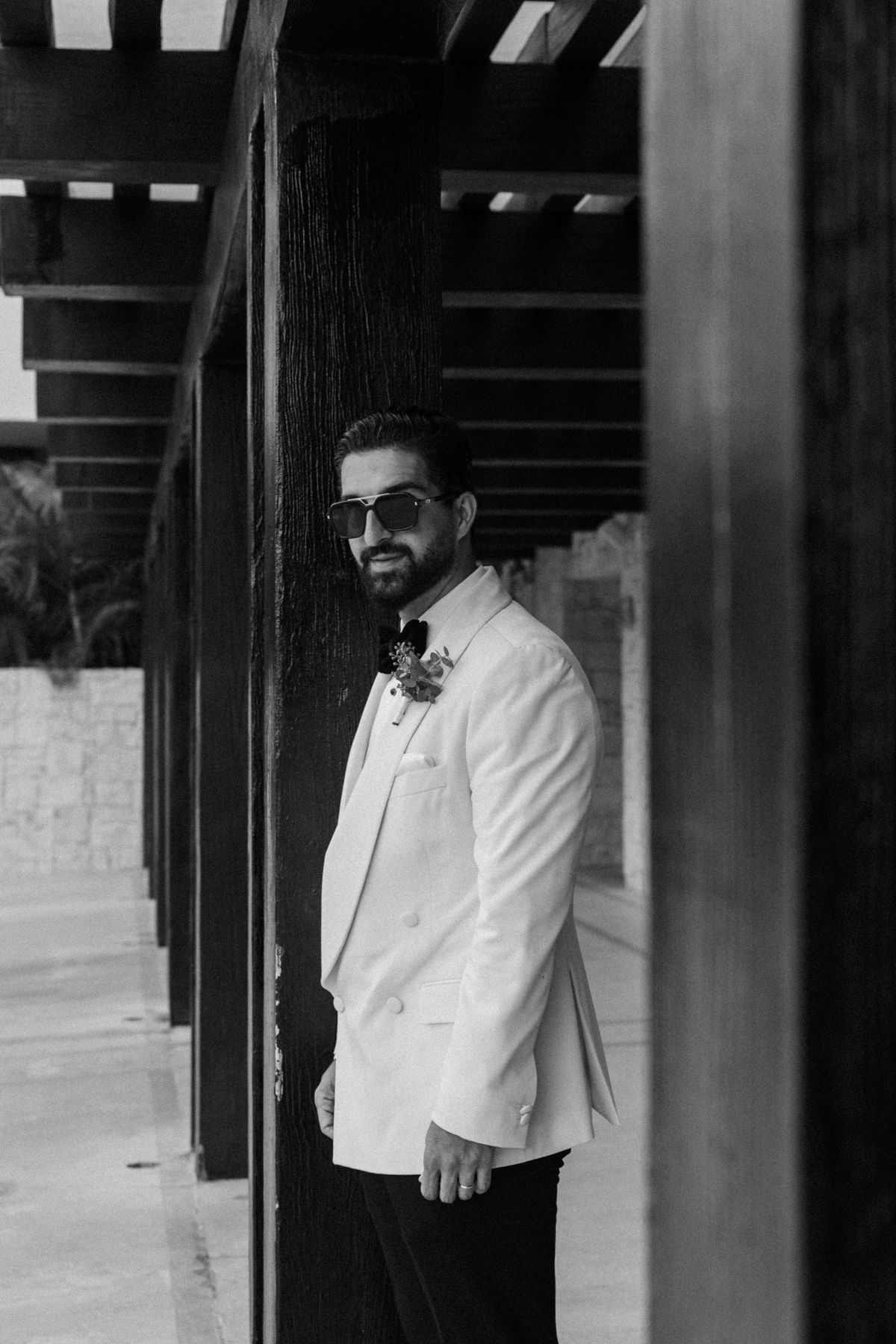 Black and white portrait of a stylish groom in white double-breasted suit with boutonniere and sunglasses.
