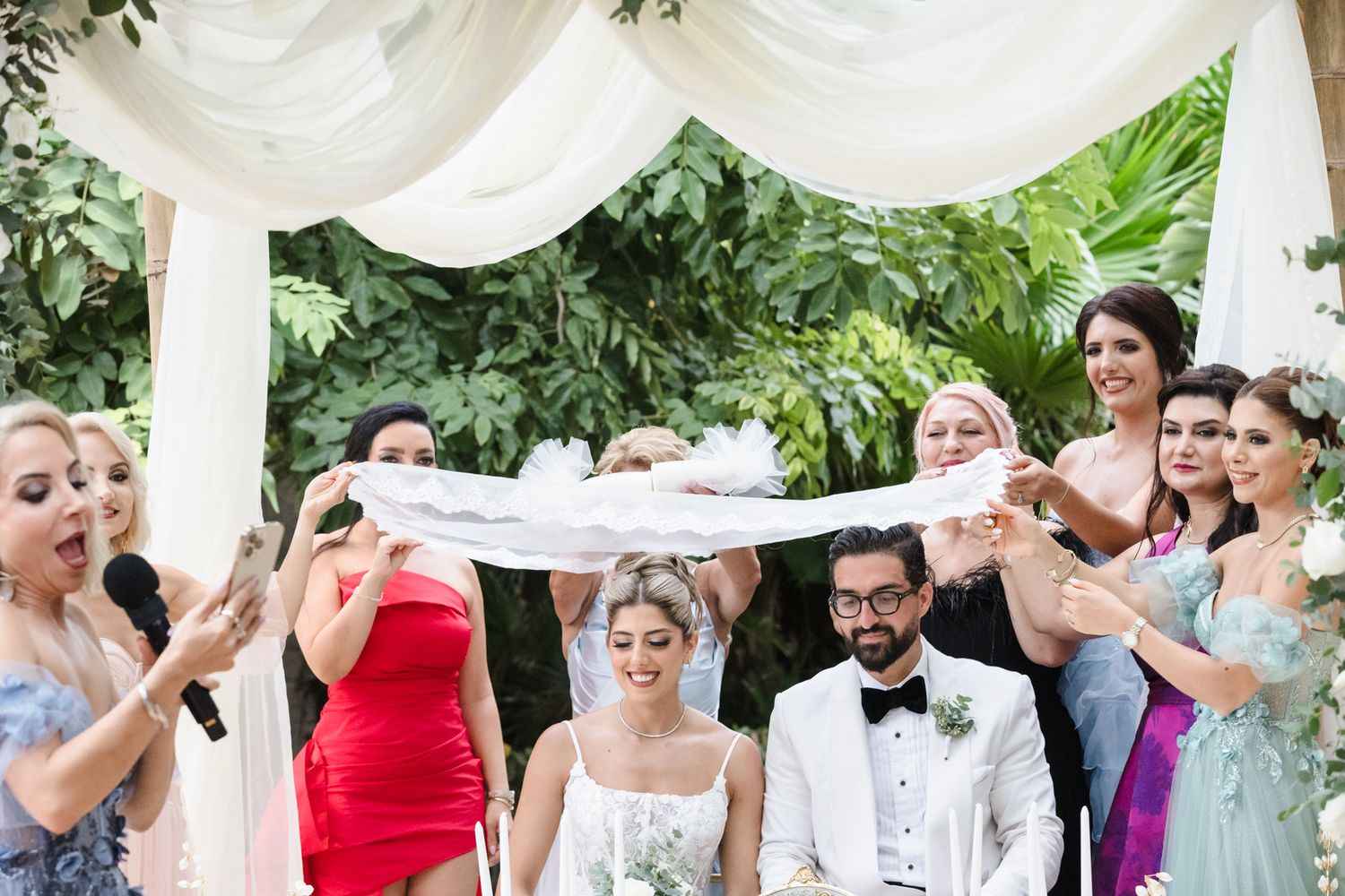 Bride and groom seated under white draped arch surrounded by bridesmaids in colorful dresses during outdoor wedding ceremony.