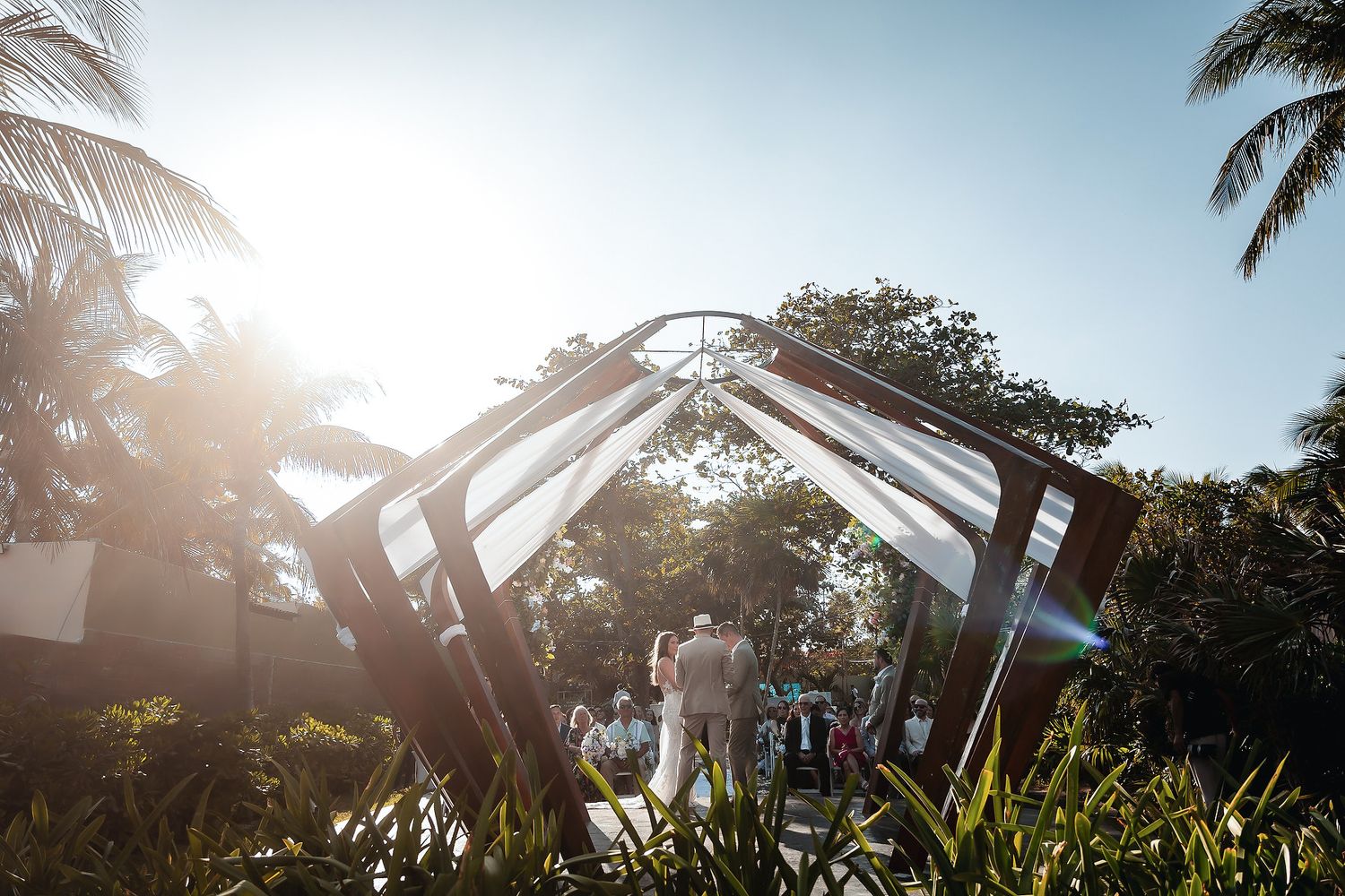 Bride and groom walk through a modern geometric arch structure surrounded by lush tropical gardens in bright sunlight.