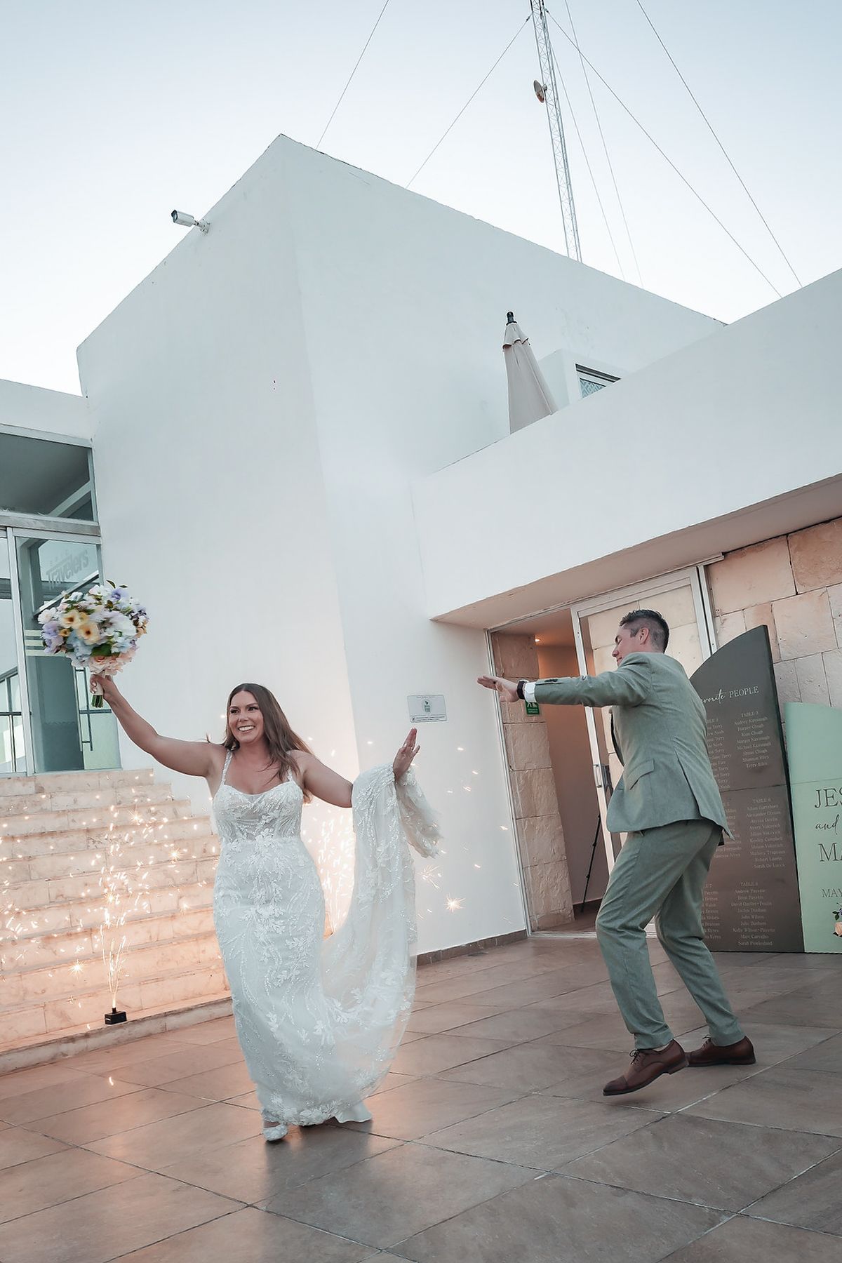 Bride in white lace gown raises bouquet while groom in sage suit celebrates outside modern white building.