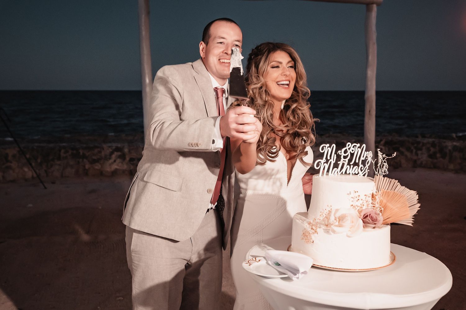 Couple cutting wedding cake on beach at night, bride in white gown and groom in tan suit, ocean backdrop.