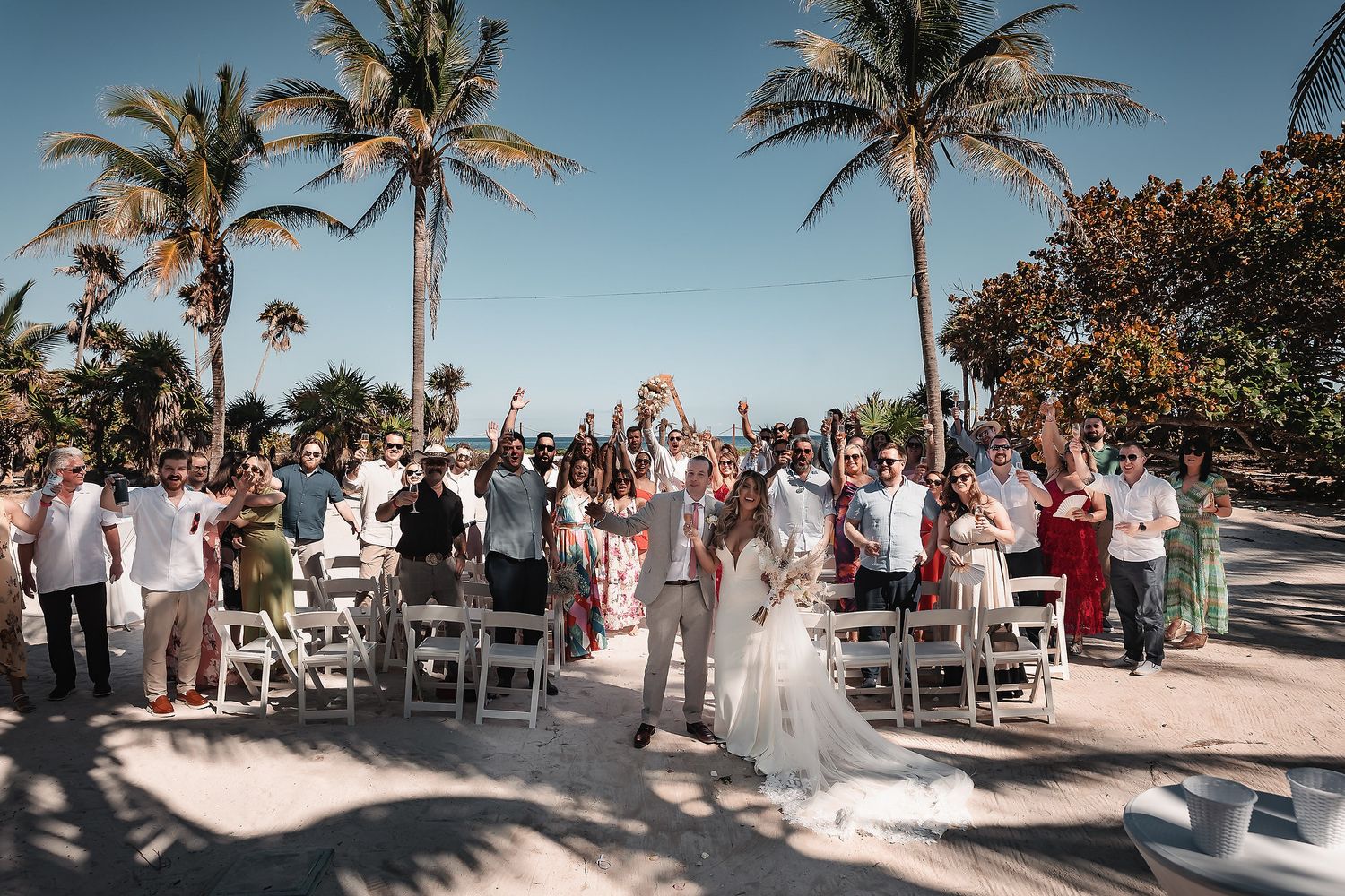 Bride and groom celebrate with guests at a tropical beach wedding ceremony surrounded by palm trees.