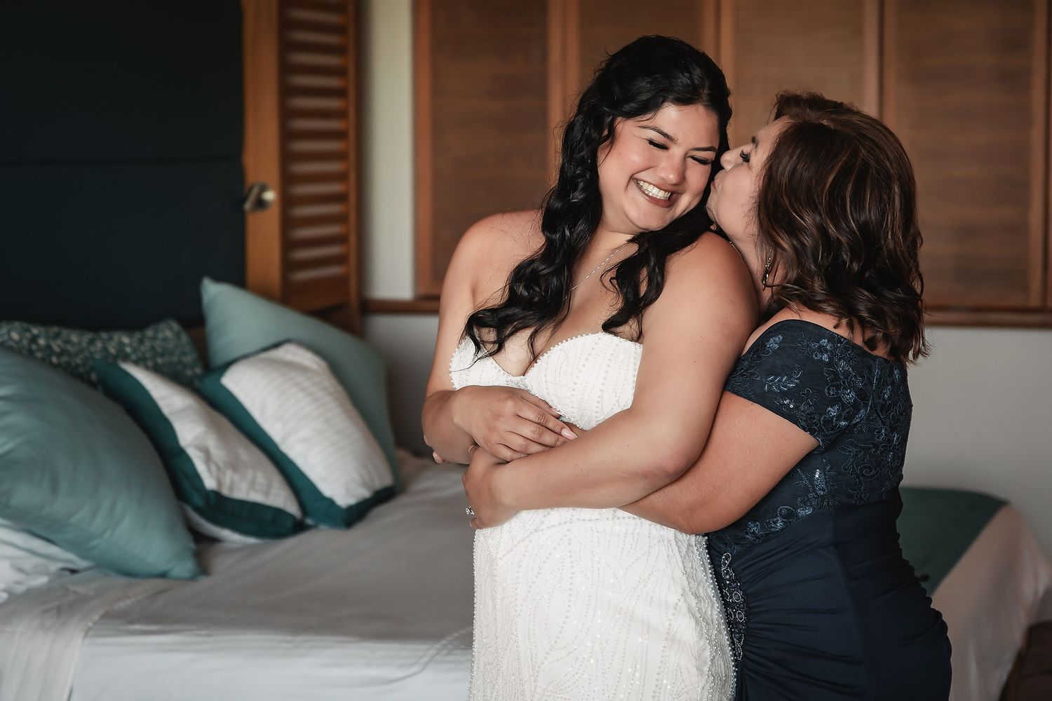 Bride in white gown smiles as woman in navy dress embraces and kisses her cheek in a beautifully decorated bedroom.