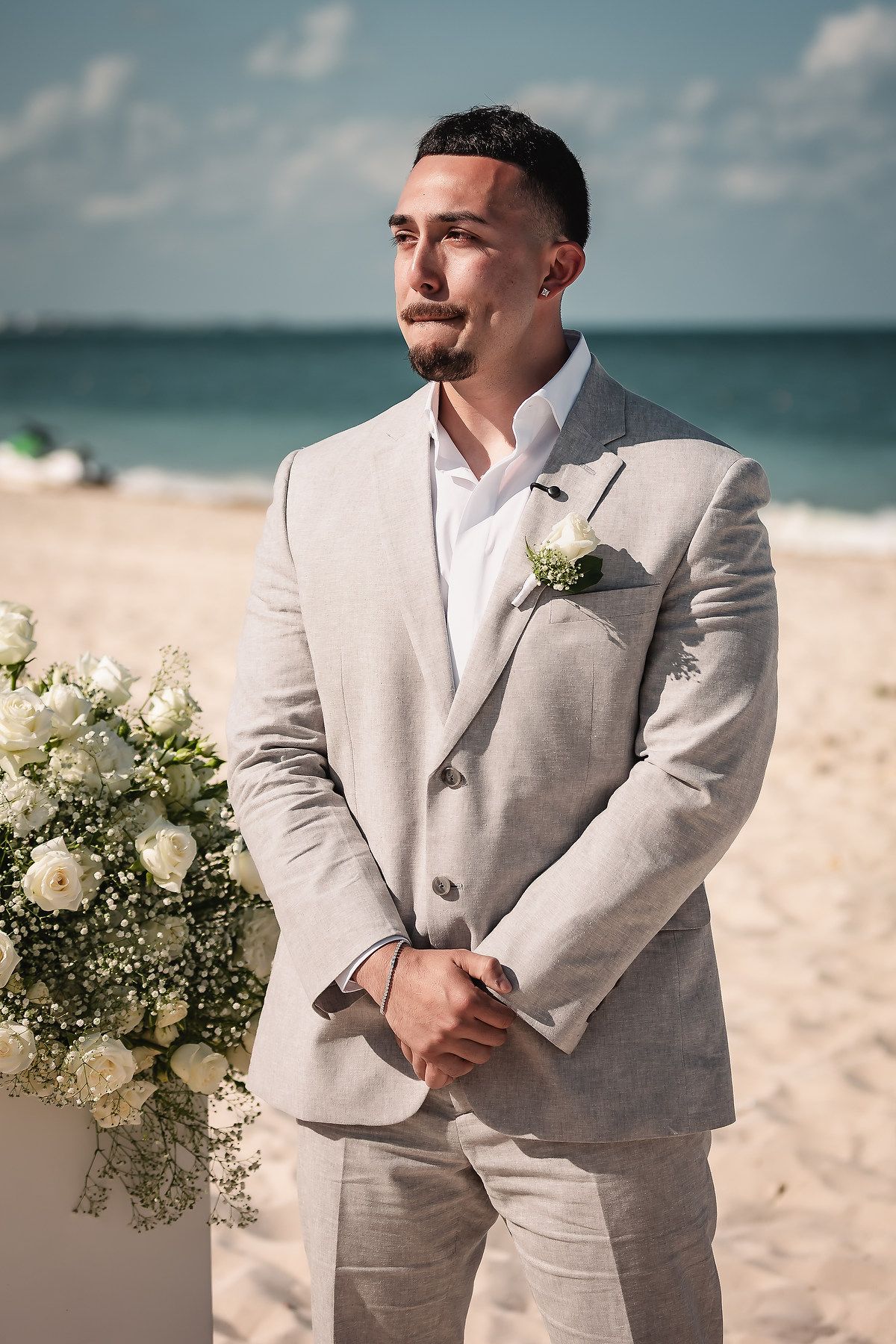 Groom in light gray linen suit with white boutonniere stands at beach wedding ceremony with ocean backdrop.