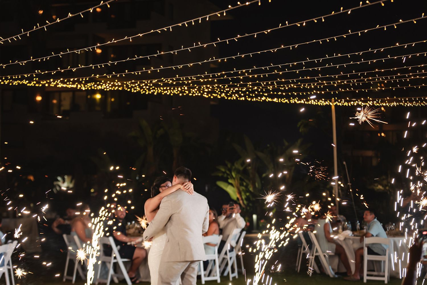 Couple shares first dance under twinkling string lights with sparklers glowing at outdoor wedding reception at night.