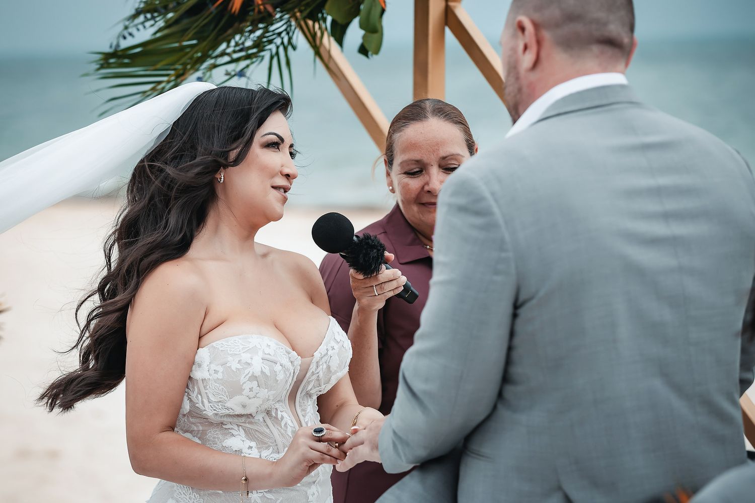 Bride reading vows at beach wedding ceremony while officiant holds microphone under tropical arch.