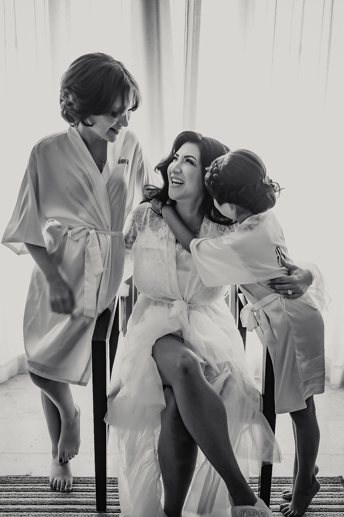Black and white photo of a bride in white robe laughing with two young girls in matching robes on wedding day.