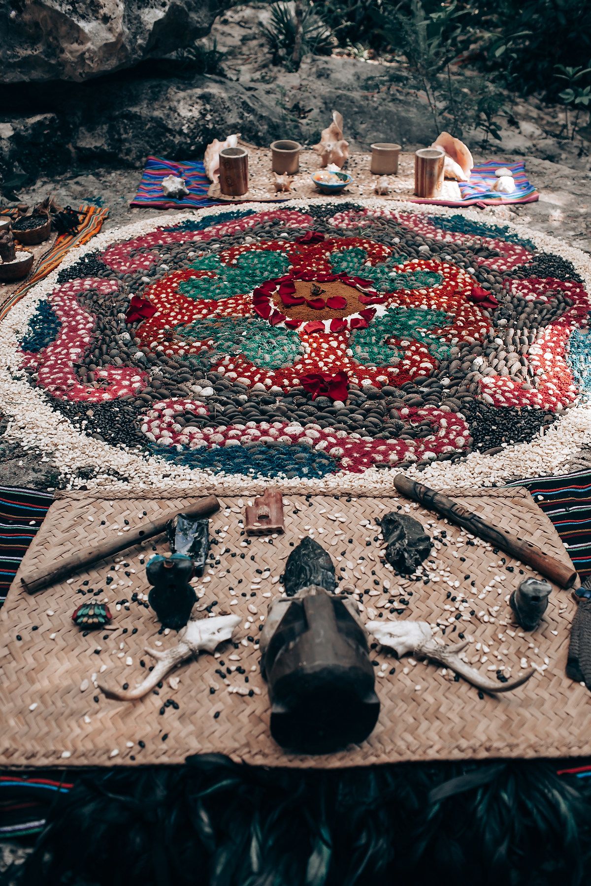 Colorful mandala made from stones and seeds on a woven mat with ritual objects and wooden cups in an outdoor ceremony.