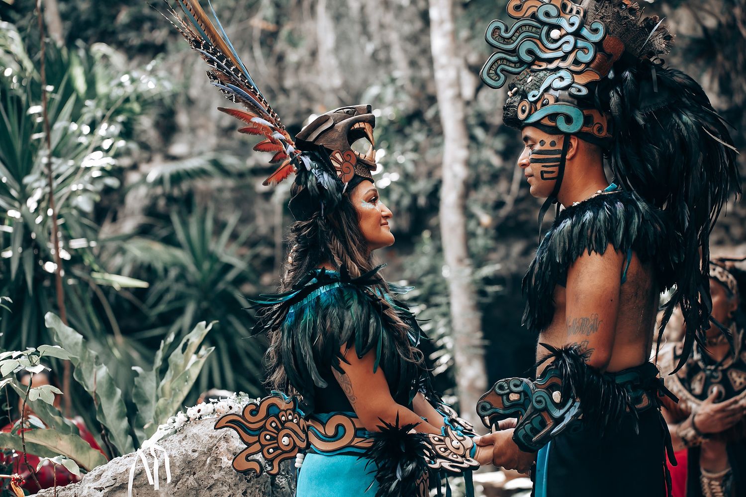 Two performers in detailed Aztec ceremonial attire with feathered headdresses facing each other in a lush forest.