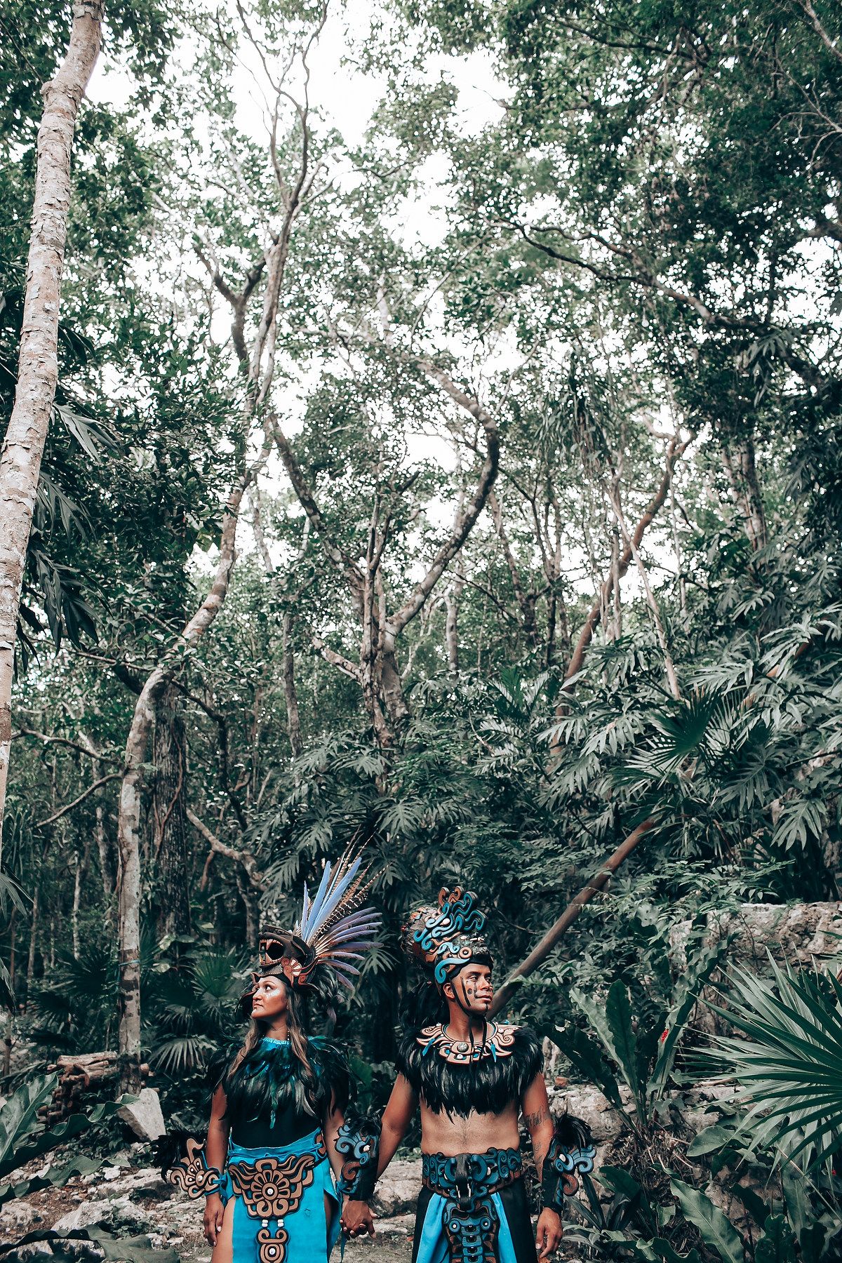 Two people in traditional Mayan warrior costumes with large feathered headdresses look up in a lush jungle.