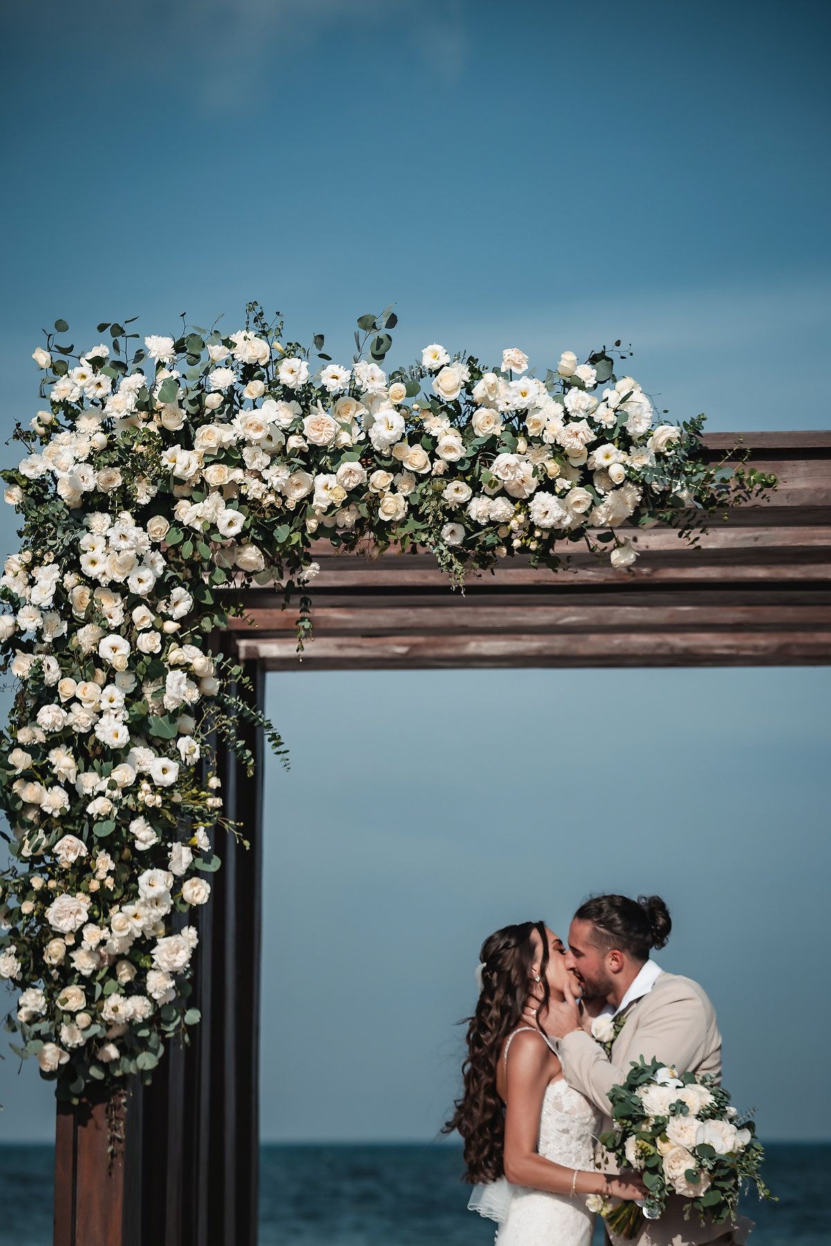 Couple kissing under a floral wedding arch adorned with white roses by the ocean on their wedding day.