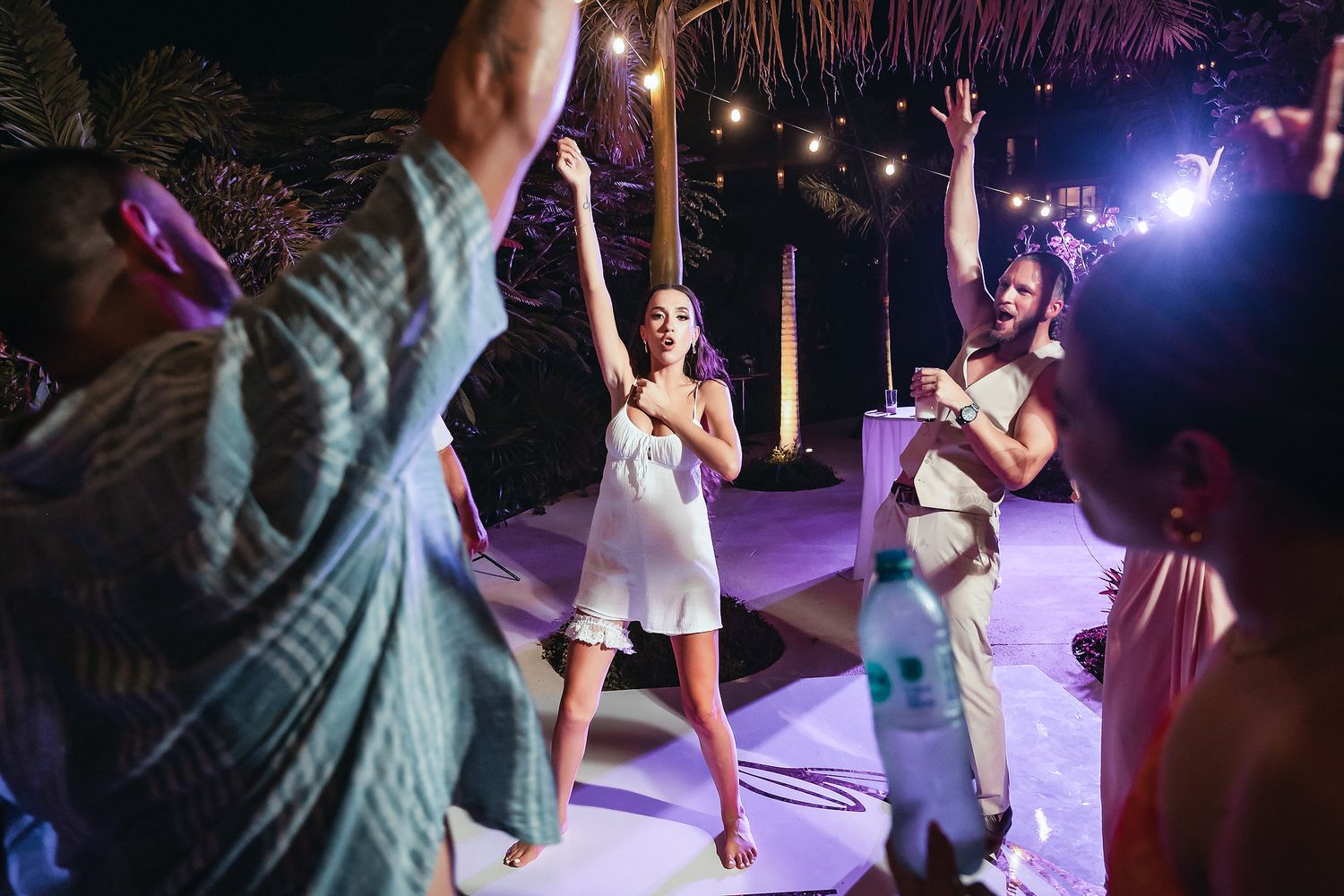 People dancing outdoors at a tropical night party with string lights and purple lighting.
