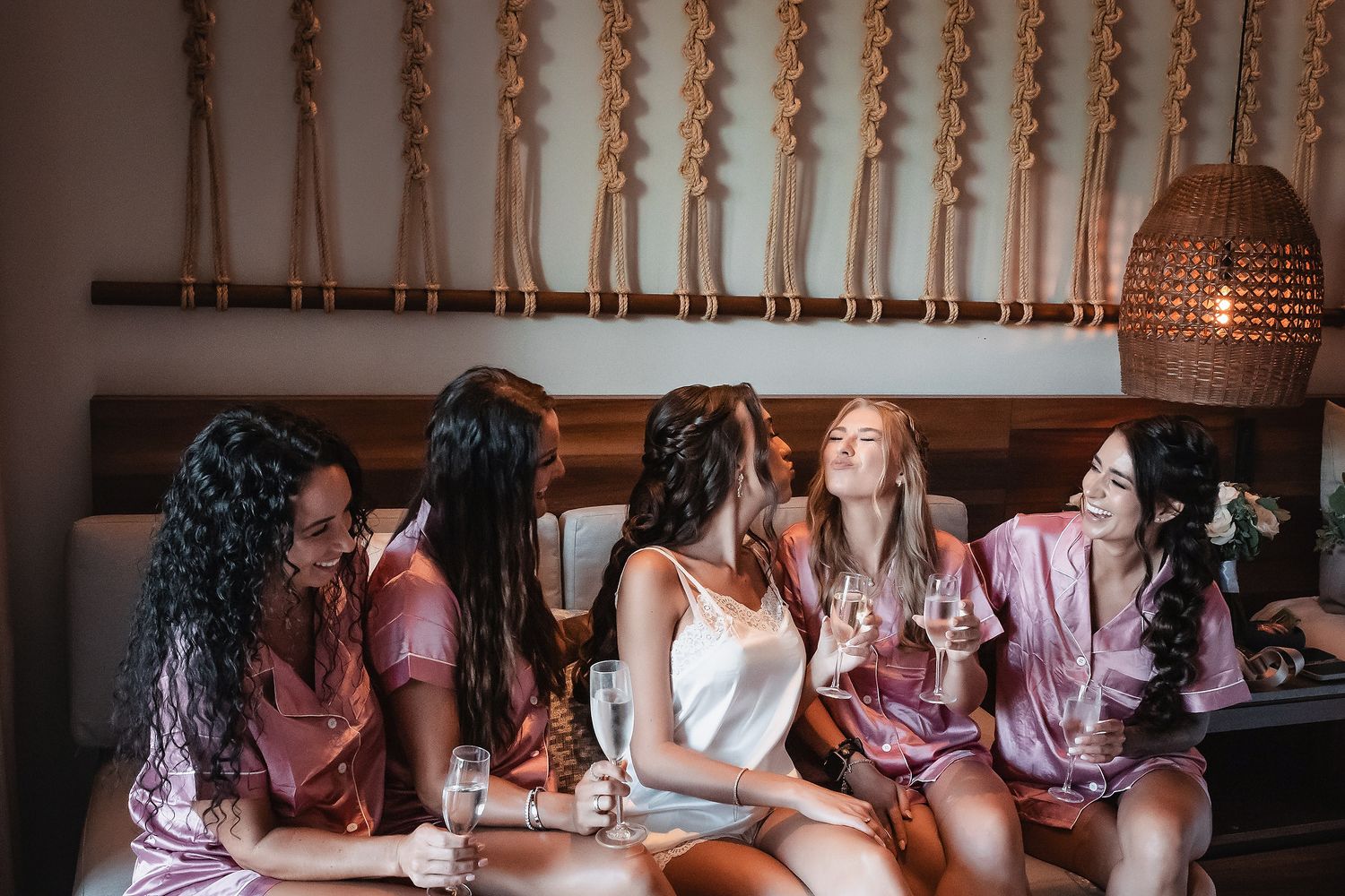 Five women in silk robes celebrate a bachelorette party, toasting with champagne glasses on a cozy bed.