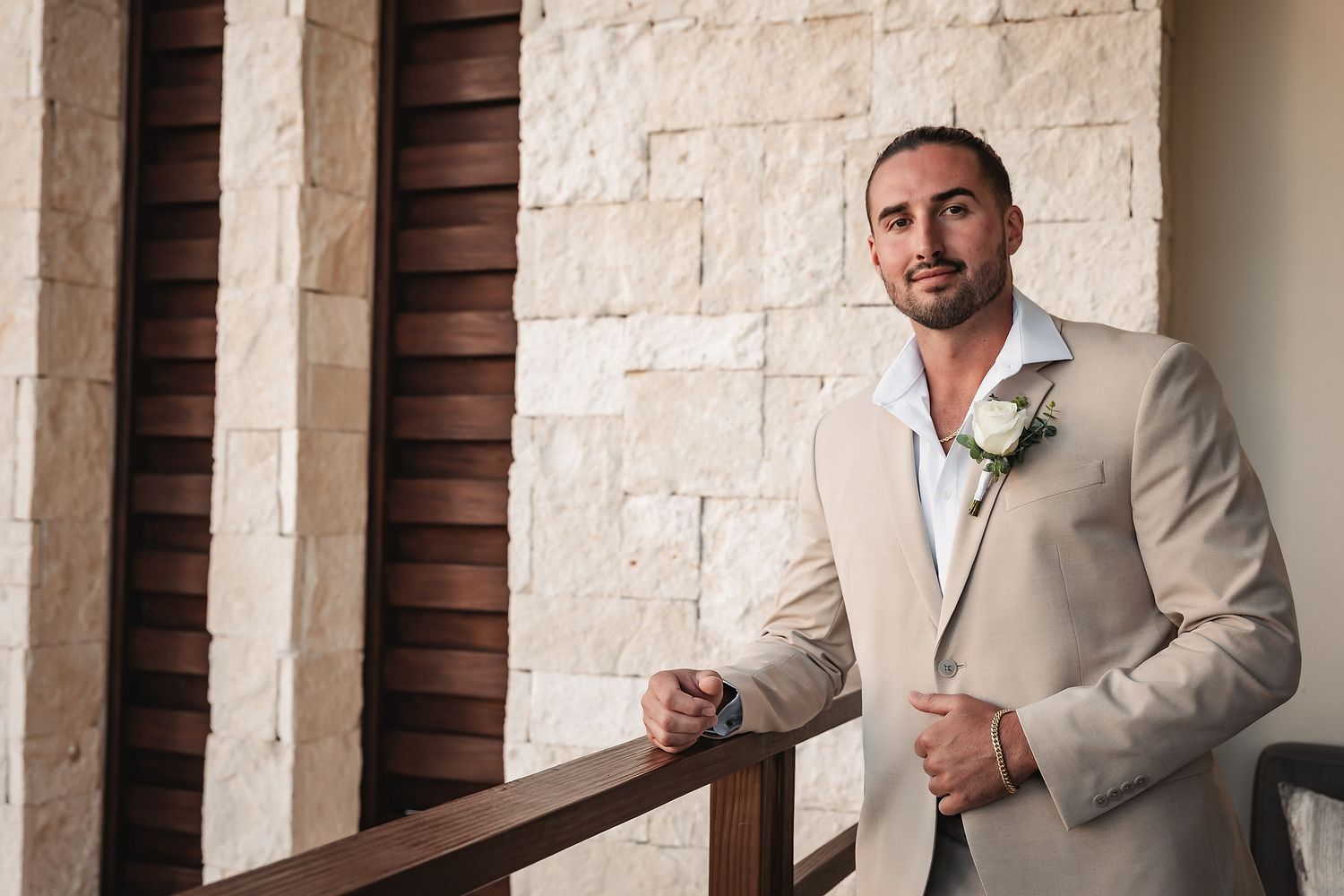 Groom in beige suit with boutonniere leaning on wooden railing against stone wall at outdoor wedding venue.