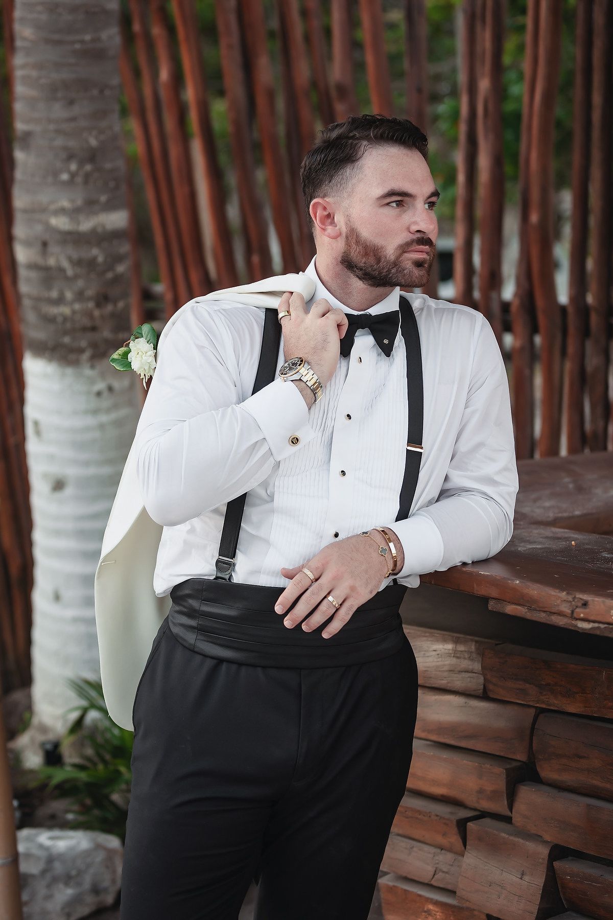 Dapper groom in black tuxedo with suspenders and bow tie holds jacket over shoulder outdoors.