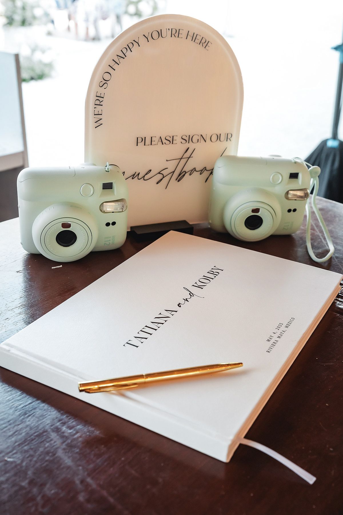 Wedding guestbook with gold pen beside mint Instax cameras and a sign reading Please Sign Our Guestbook on a wooden table.