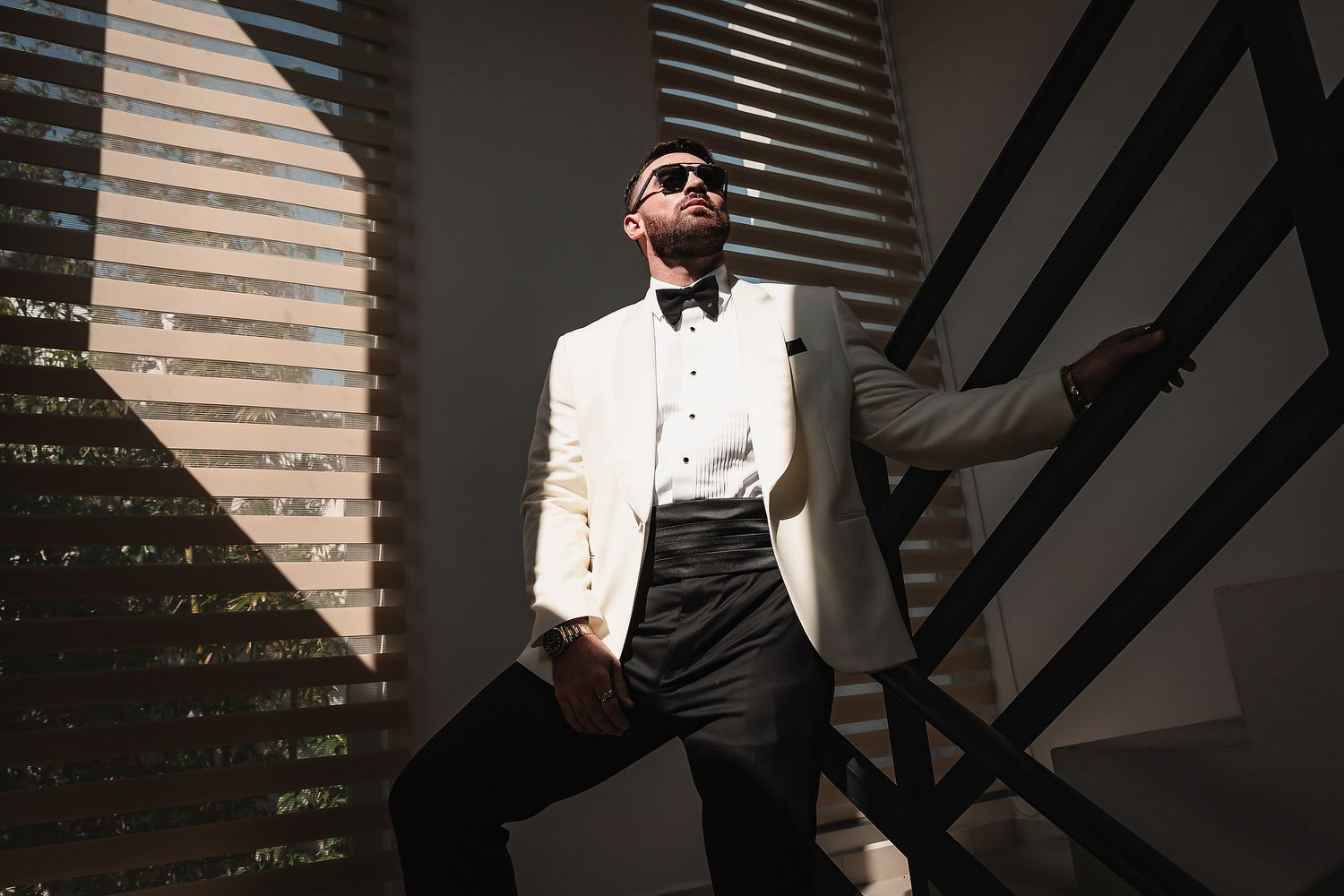 A stylish man in a white tuxedo jacket and black bow tie poses confidently wearing sunglasses in dramatic light.