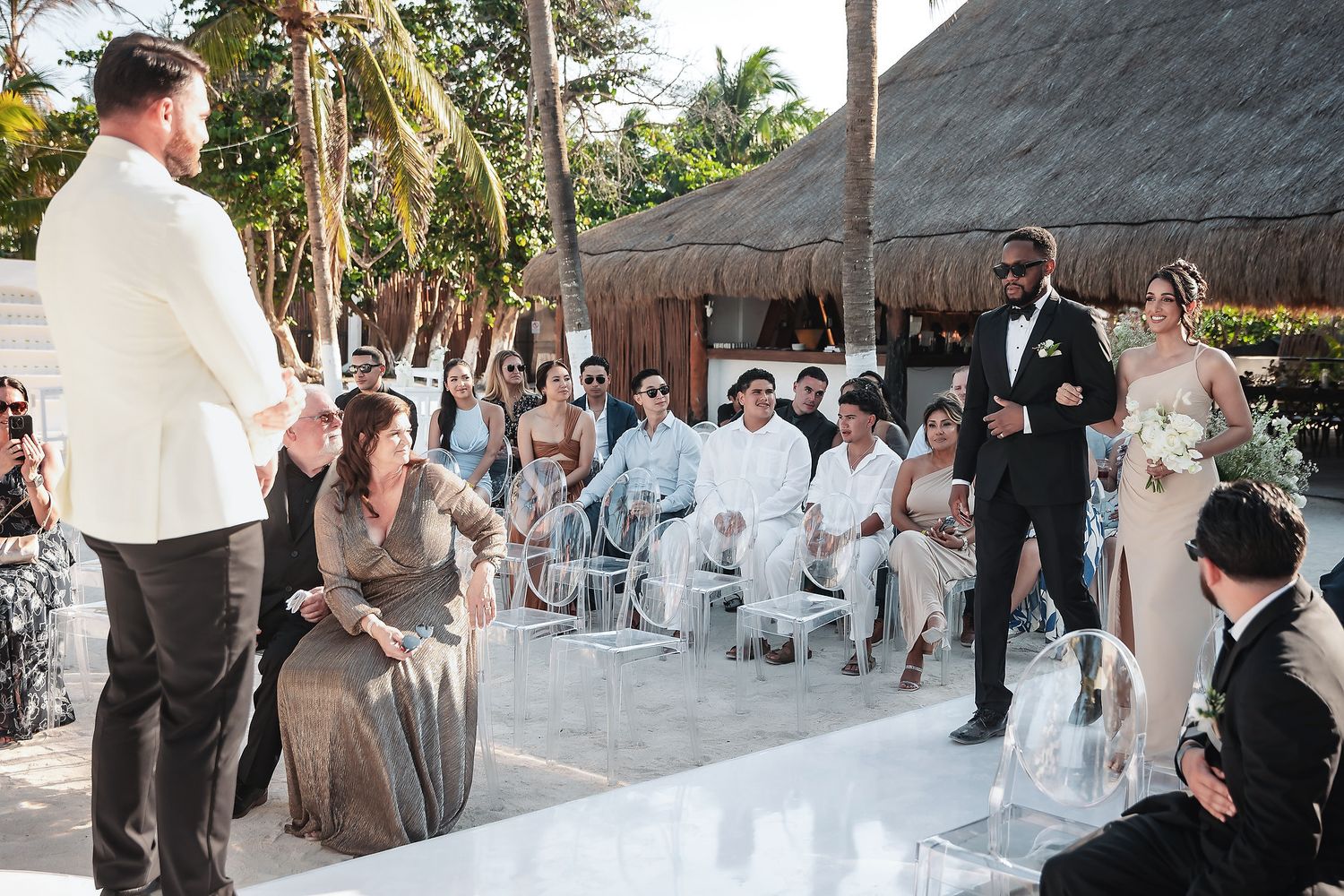 Outdoor wedding ceremony with guests seated, groom in white jacket, bride in elegant gown, tropical venue with thatched roof.