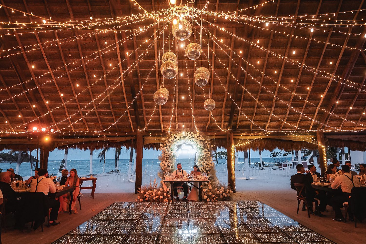 Romantic outdoor wedding reception under string lights and disco balls at a beachside venue with floral decor.
