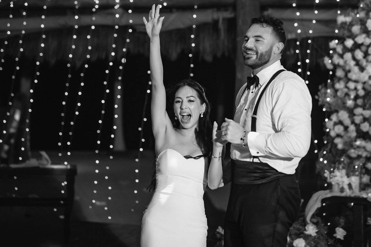 Black and white photo of a joyful bride and groom celebrating at their wedding reception with twinkling lights backdrop.