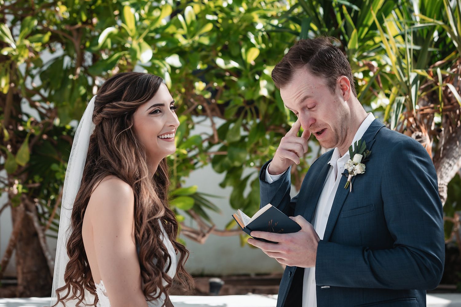Emotional groom reads wedding vows while smiling bride listens during outdoor beach ceremony surrounded by tropical greenery.