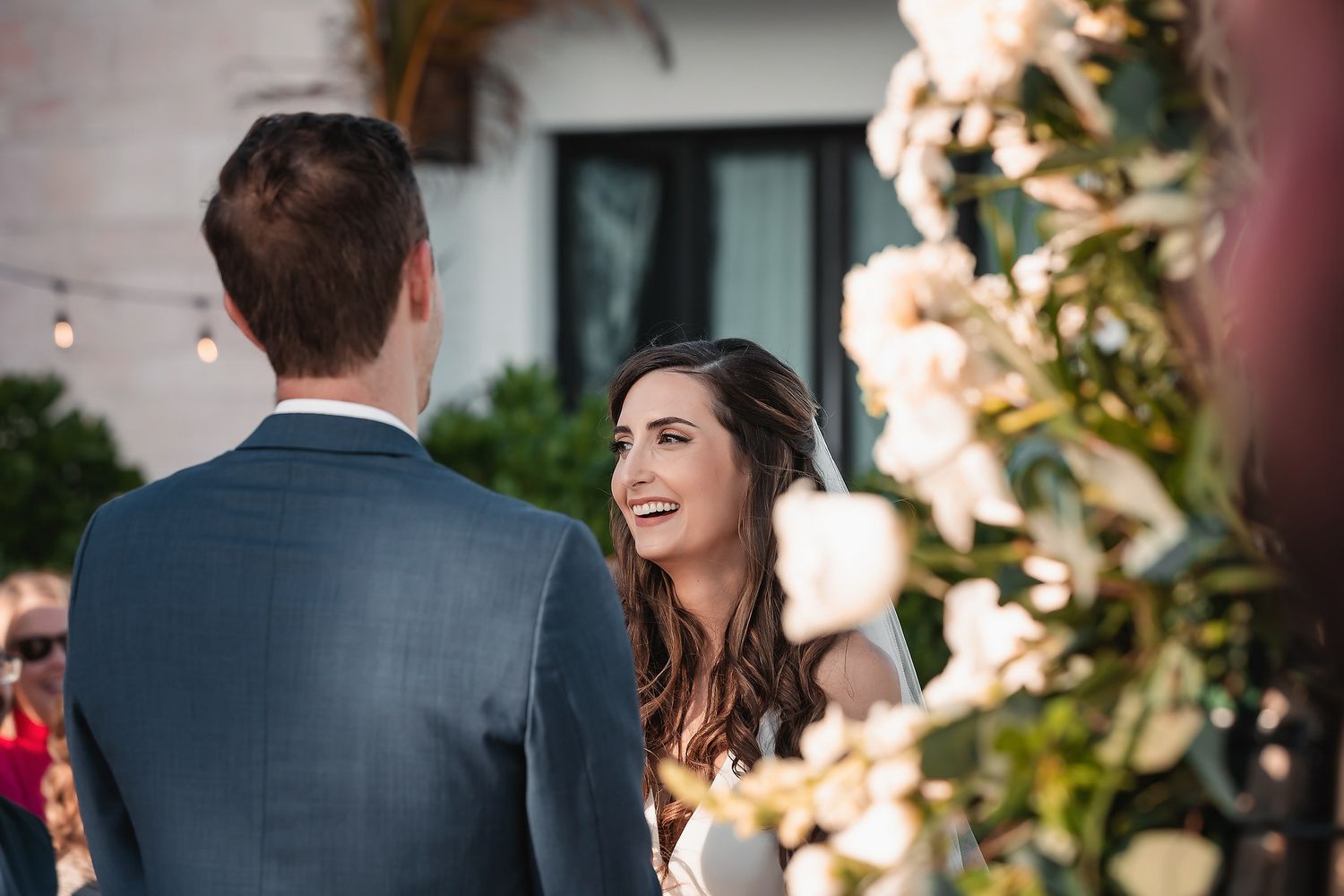 Bride smiling joyfully at groom during outdoor wedding ceremony with white floral arch and string lights.