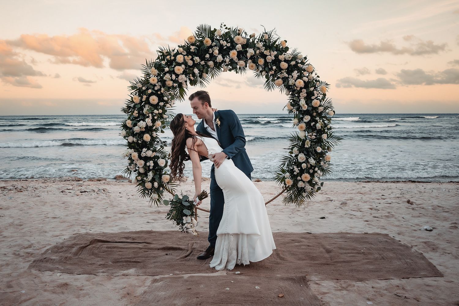Couple sharing a kiss under a circular white floral arch on a sandy beach at sunset.