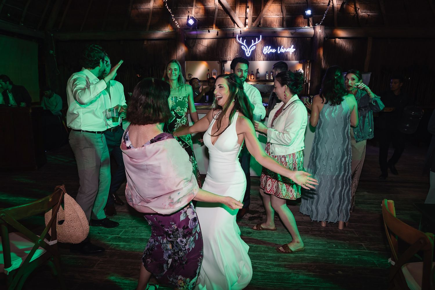 Bride in white dress dancing joyfully with guests at a vibrant wedding reception under colorful lights.
