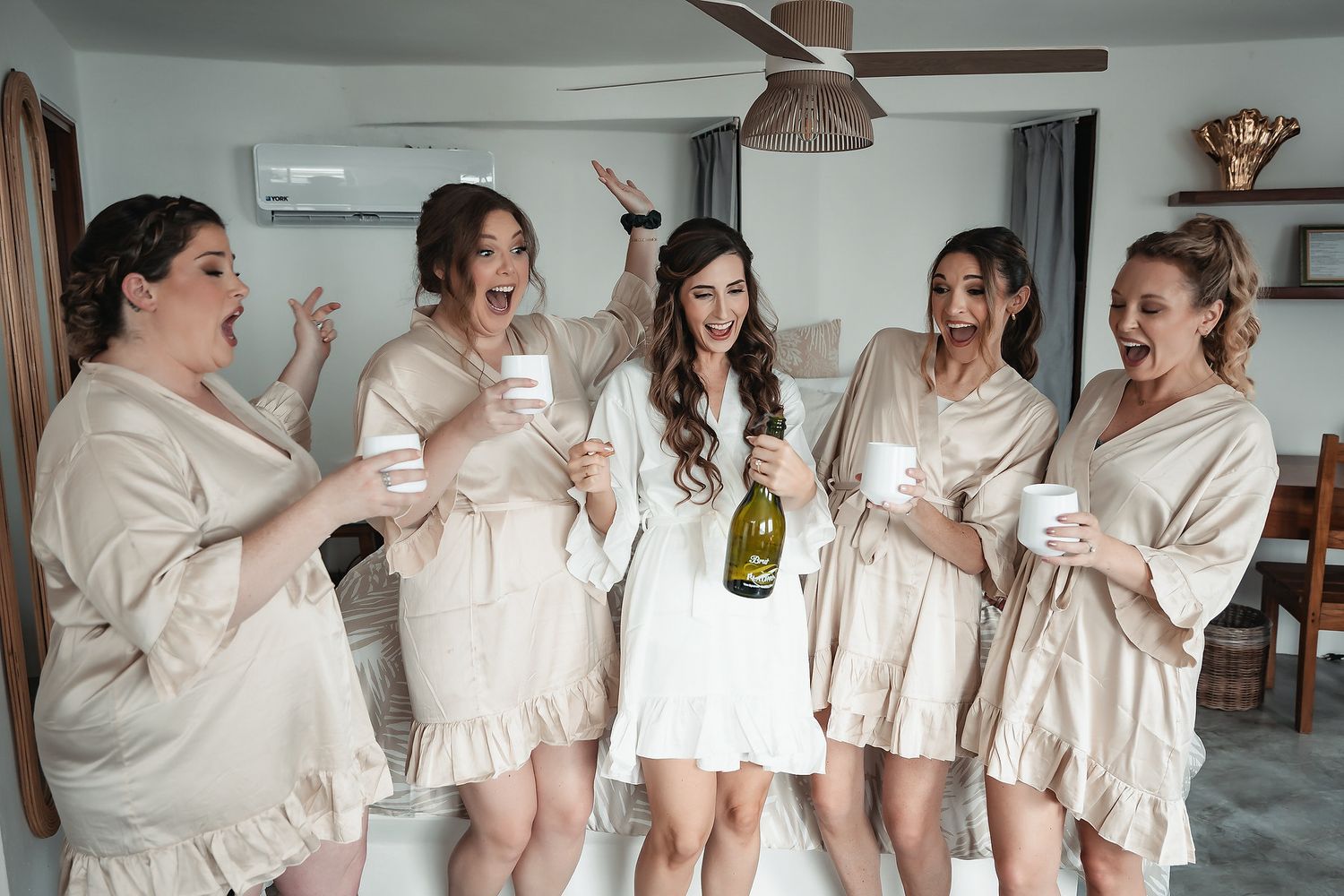 Bride in white dress celebrates with bridesmaids in champagne robes, holding wine glasses and champagne bottle indoors.