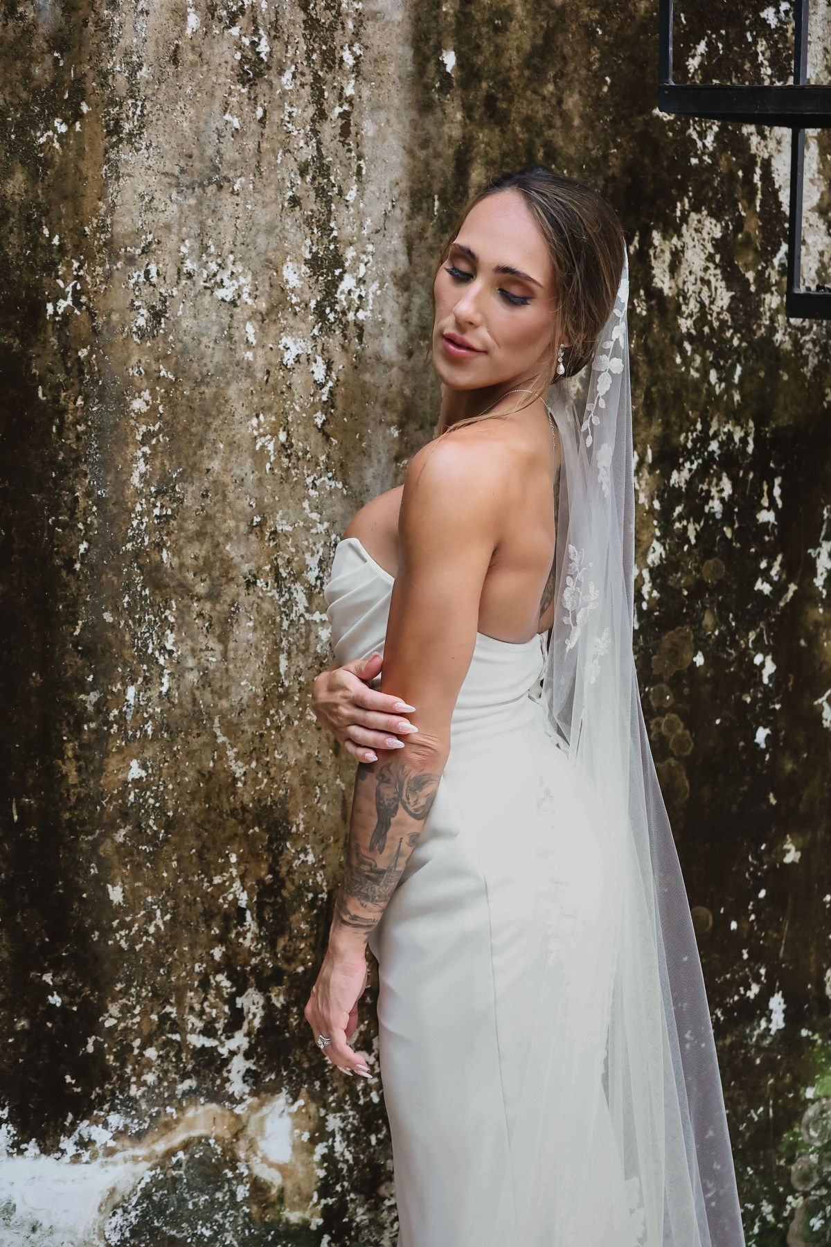 Bride in strapless white gown and veil with arm tattoo poses elegantly against a mossy stone wall.