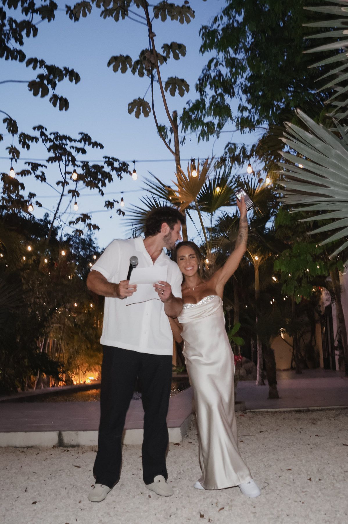 Bride in satin gown and groom in white shirt celebrate outdoors at night under string lights at tropical wedding reception.