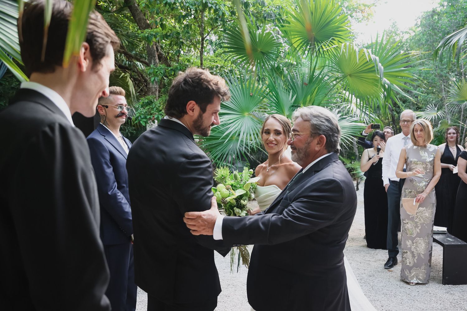 Groom shaking hands with older man at elegant outdoor wedding reception surrounded by guests.