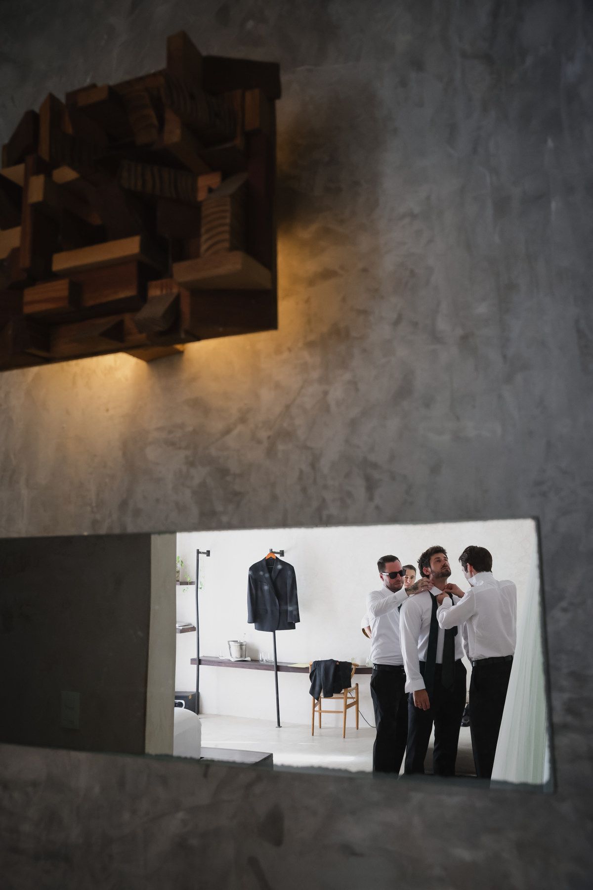 Mirror reflection of three men in white shirts adjusting ties in a modern room with a suit on display.