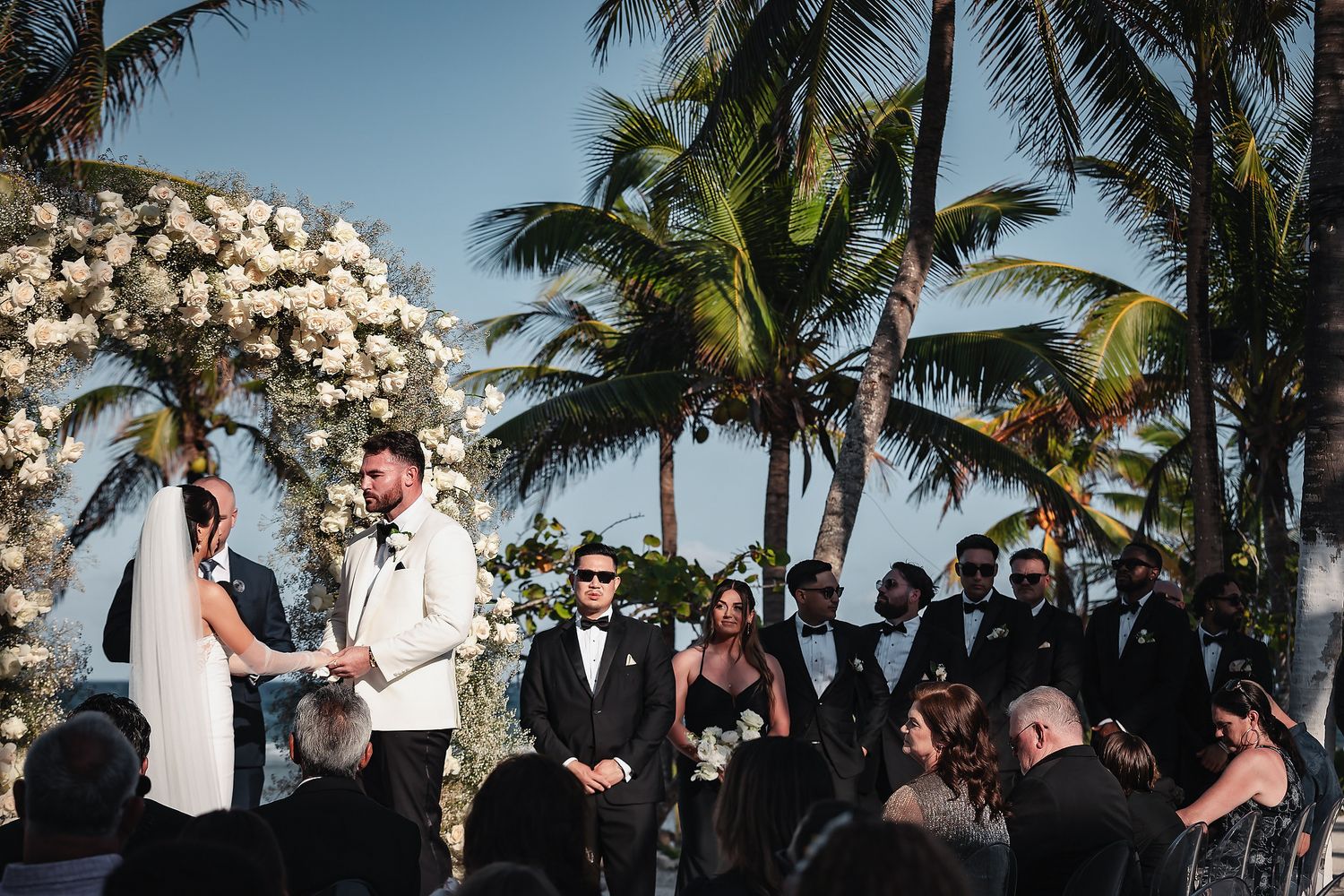 Outdoor beach wedding ceremony with couple exchanging vows under floral arch surrounded by palm trees and guests.