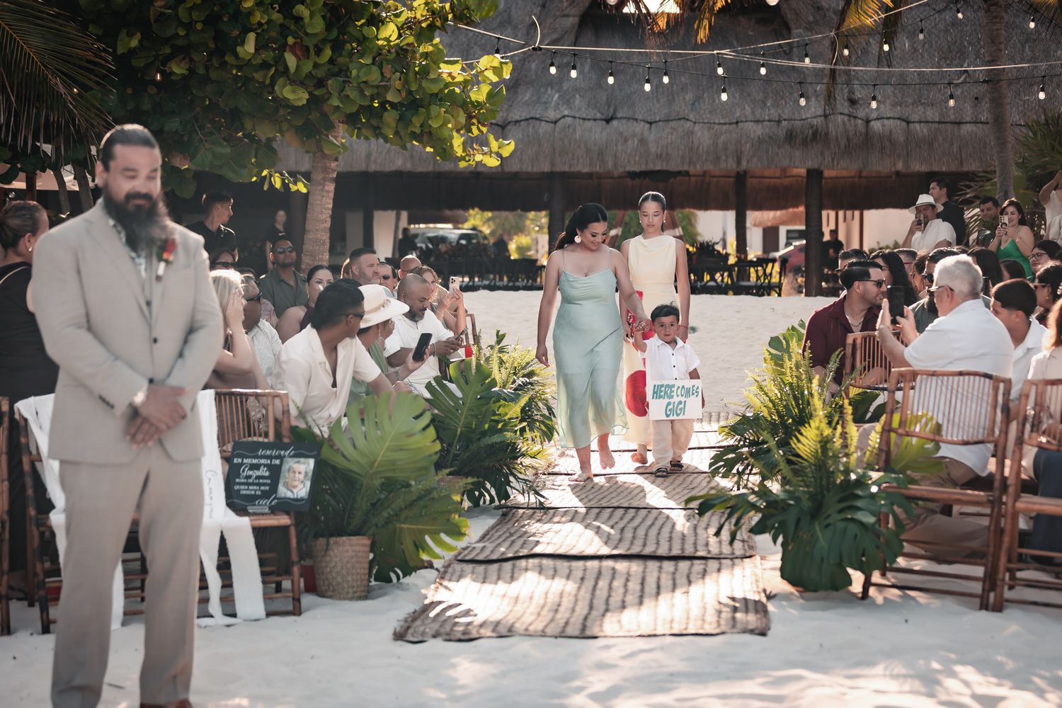 Outdoor beach wedding ceremony with guests seated on wooden benches, tropical plants, and string lights at a coastal venue.