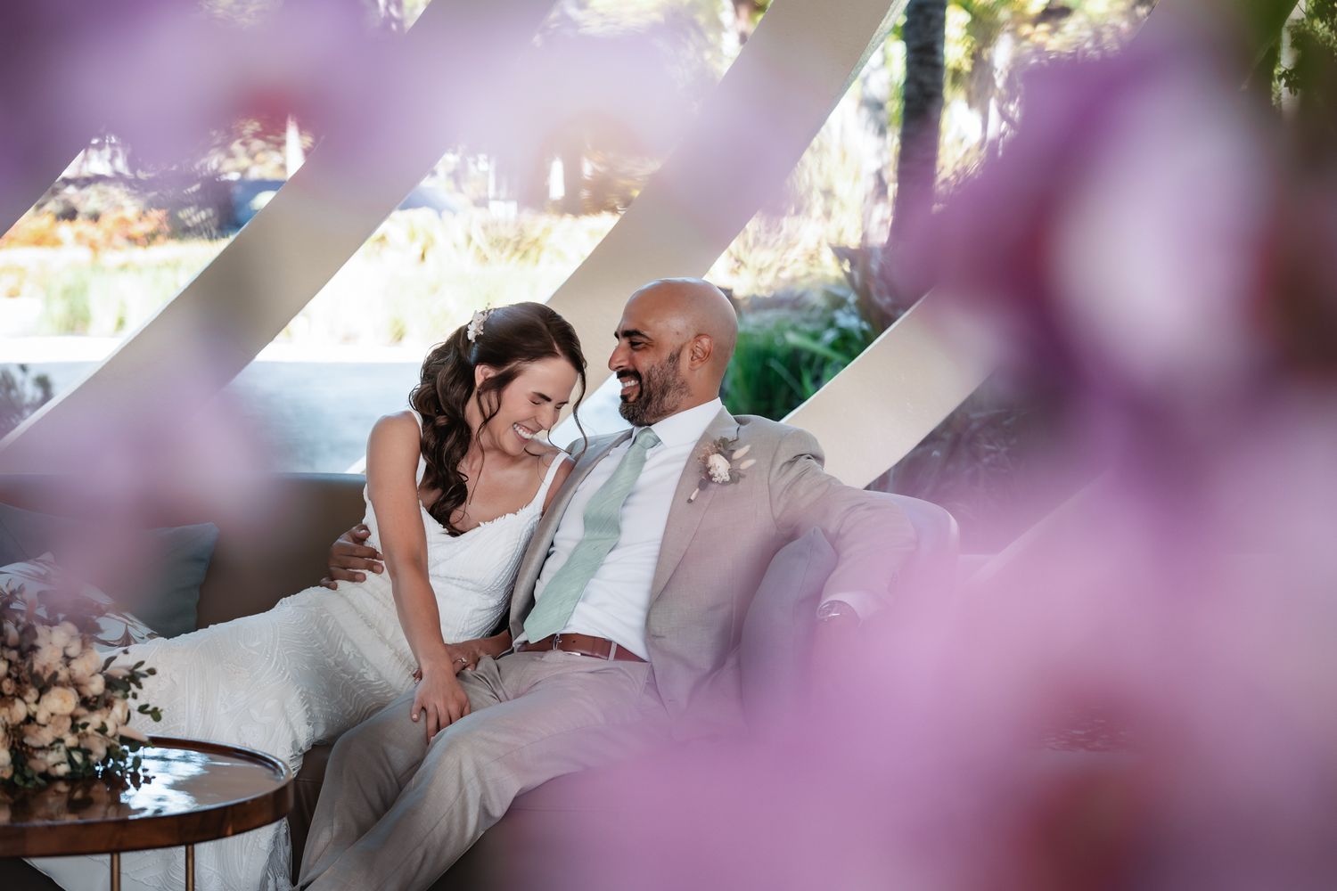 Bride and groom sitting close together on a couch, framed by purple blossoms at an outdoor wedding venue.