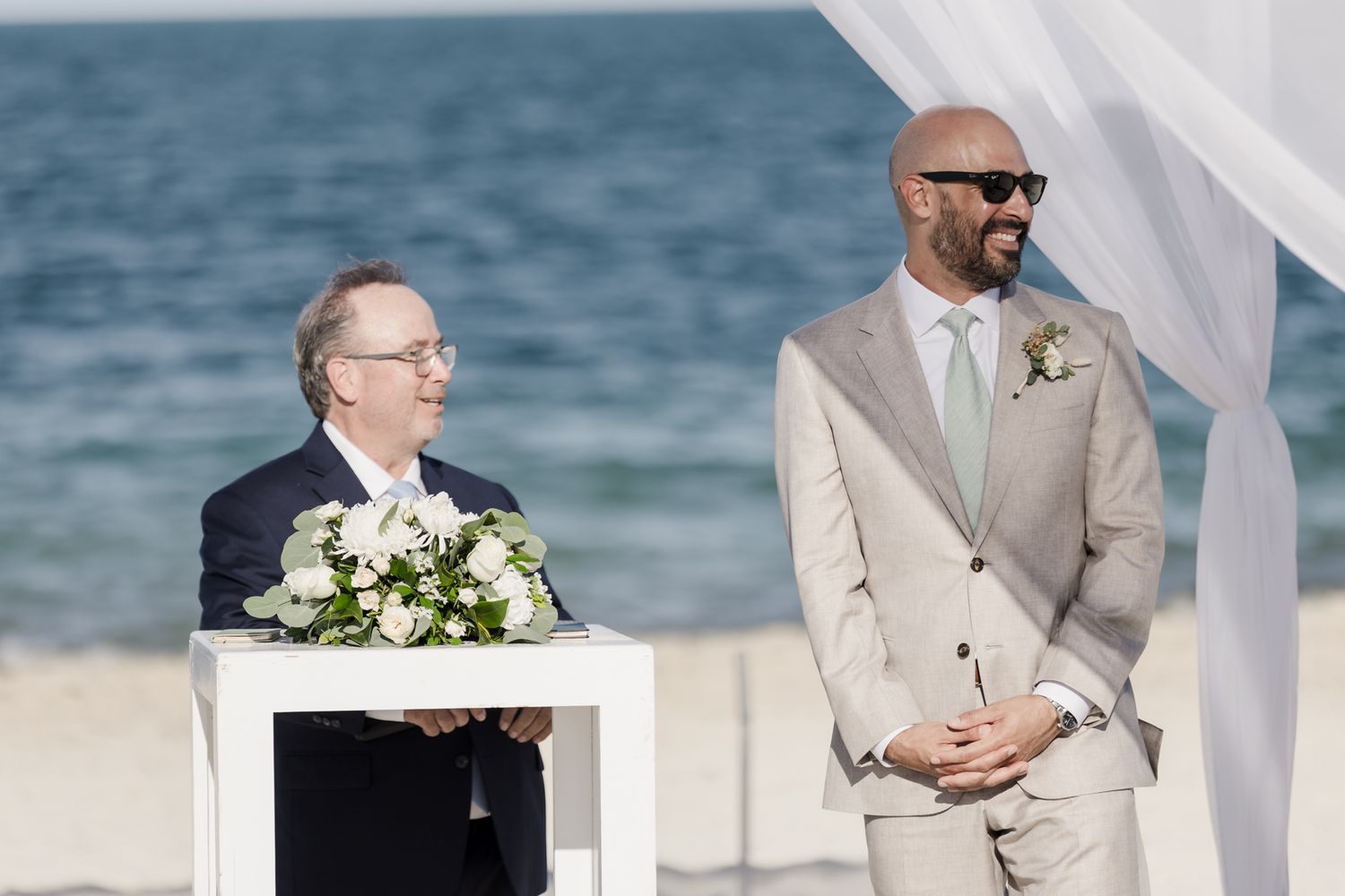 Groom in beige suit stands at beach wedding altar beside officiant holding white floral bouquet with ocean backdrop.