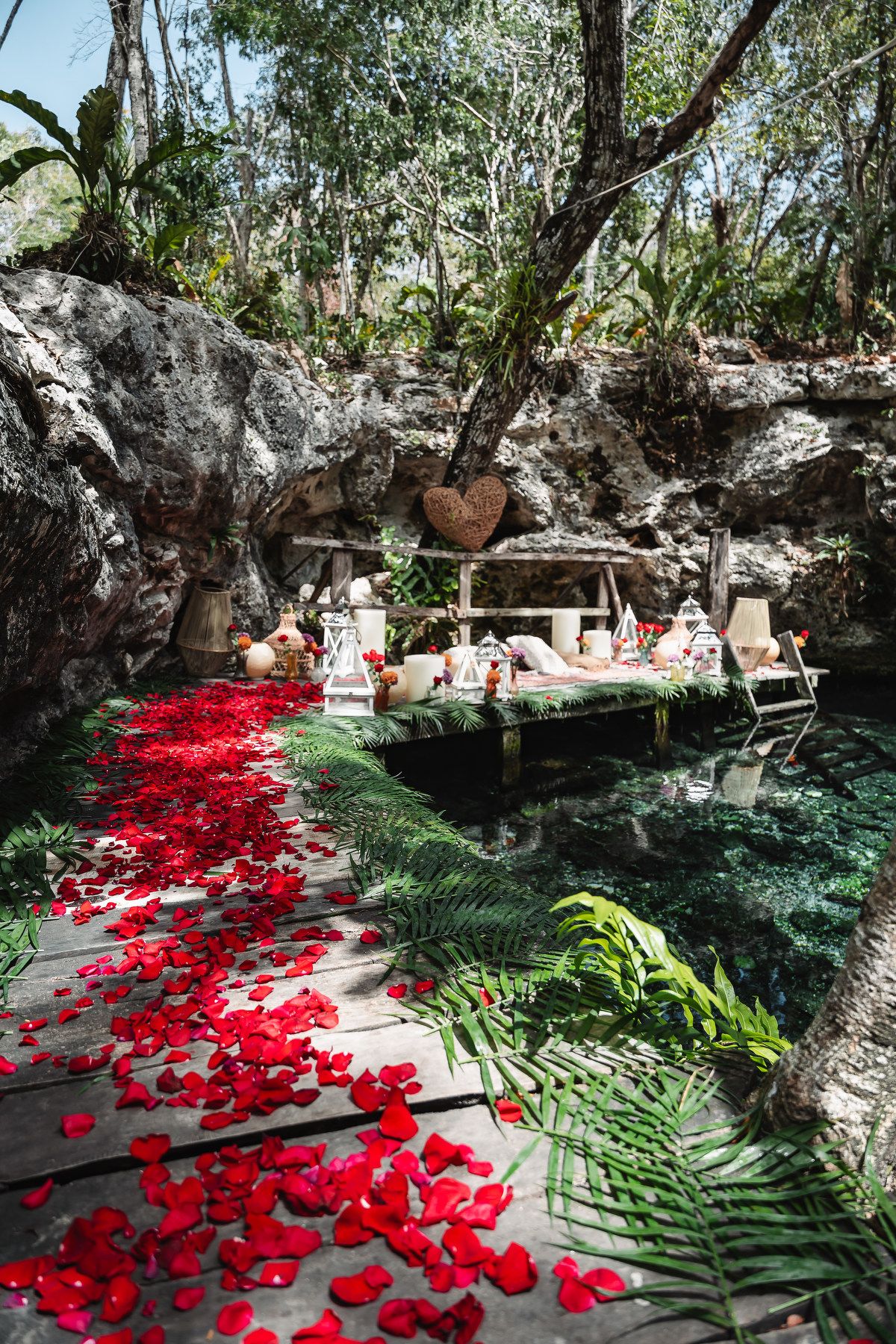 Romantic jungle cenote setup with red rose petals on wooden deck, heart decor, lanterns, and lush tropical greenery.
