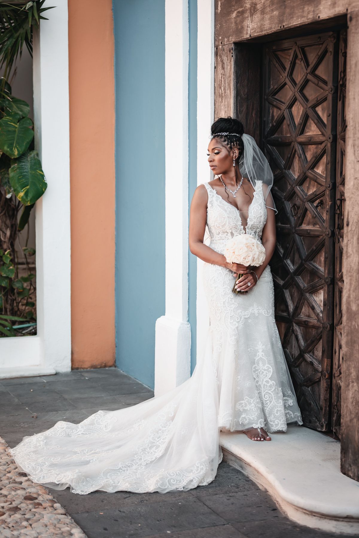 Elegant bride in lace wedding gown with veil holds bouquet beside ornate wooden door with colorful colonial architecture.