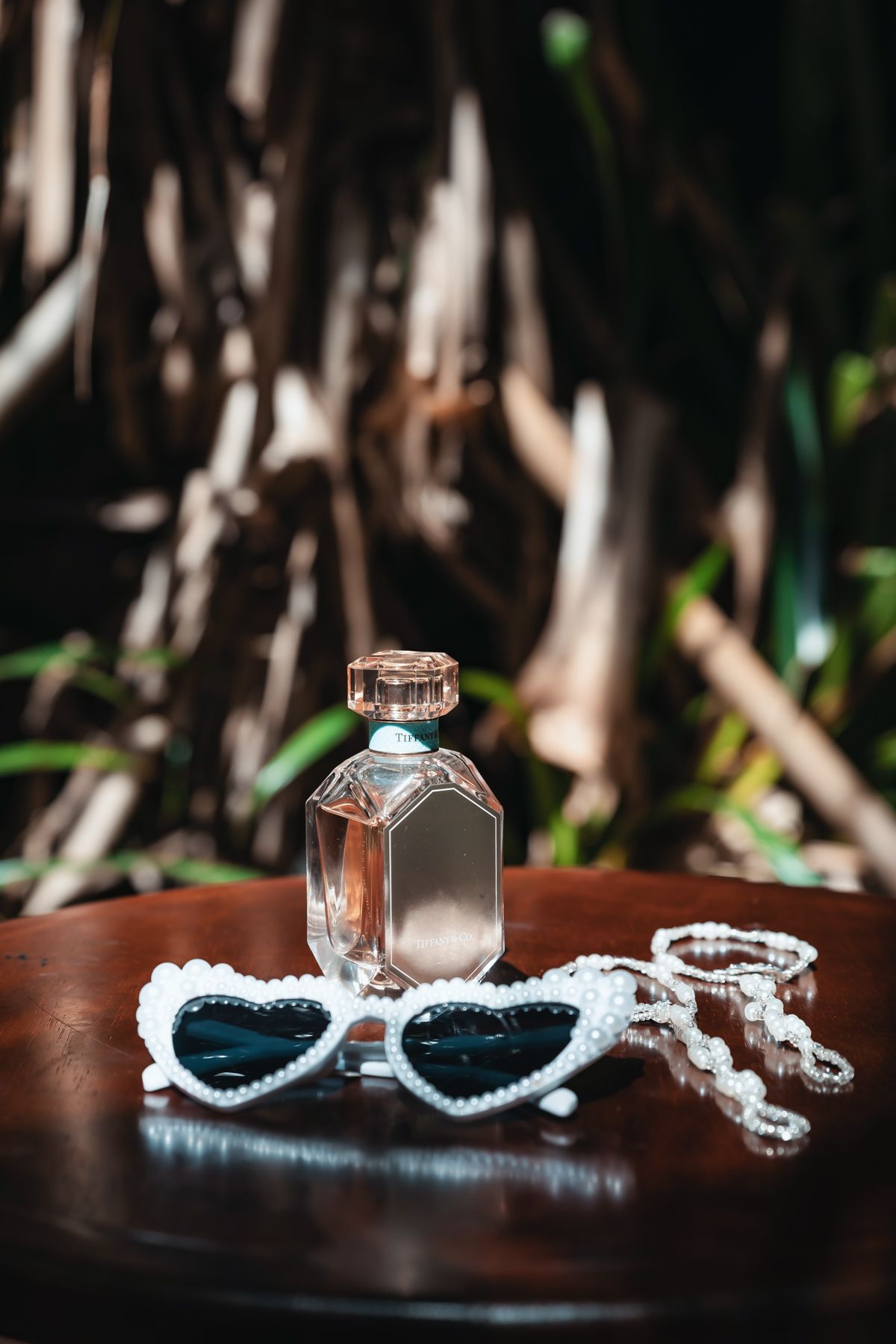 Heart-shaped pearl sunglasses and pearl necklace beside a glass perfume bottle on a wooden table outdoors.