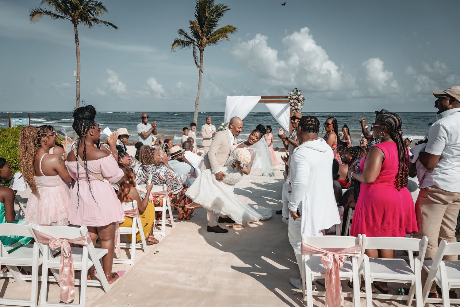 Outdoor beach wedding ceremony with guests seated in white chairs, couple at altar with ocean and palm trees in background.