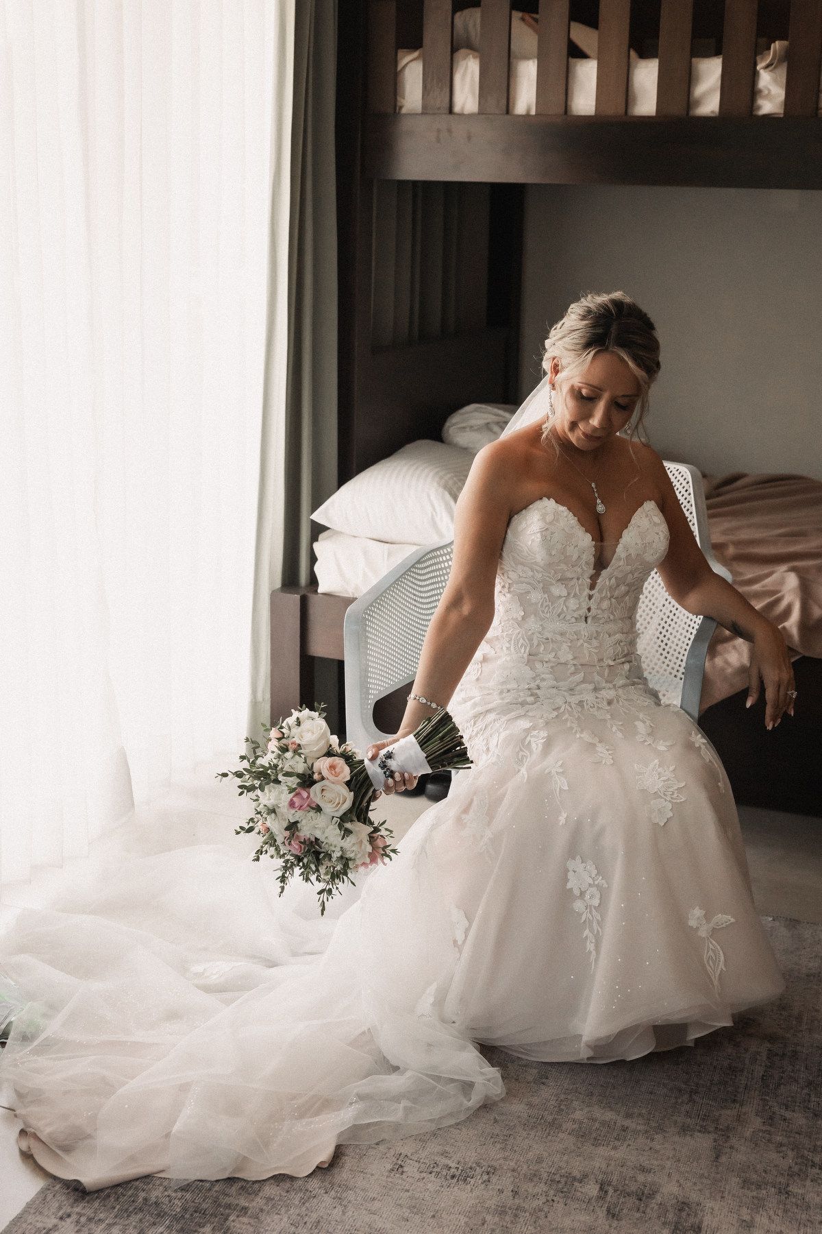 Bride in strapless lace mermaid wedding gown holding bouquet of pink roses in hotel room.