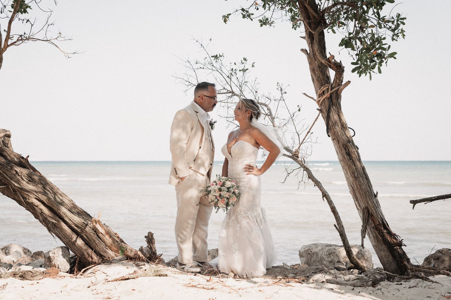Bride and groom pose by weathered tree on sandy beach with ocean backdrop.