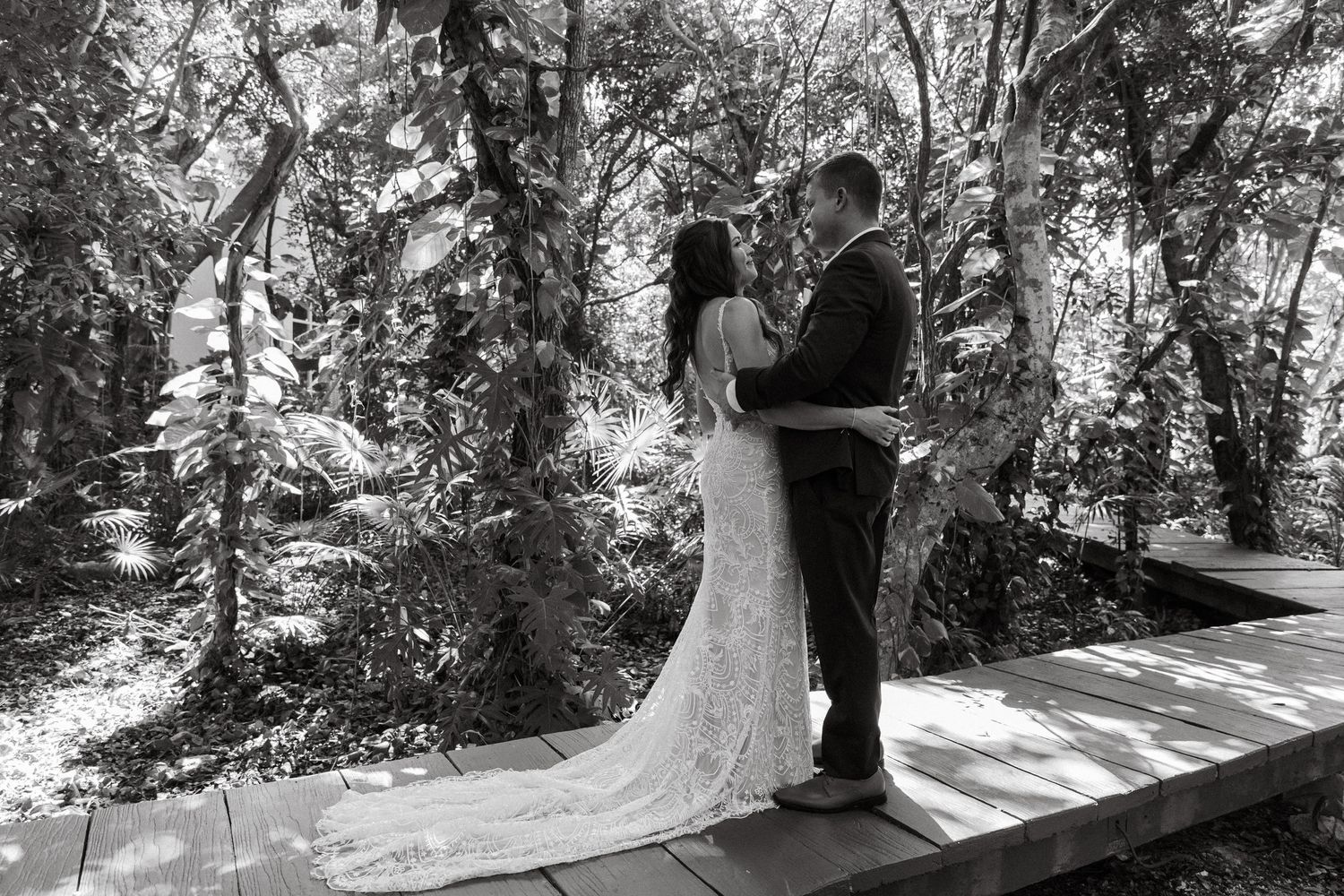 Black and white photo of bride and groom embracing on wooden bridge in forest.