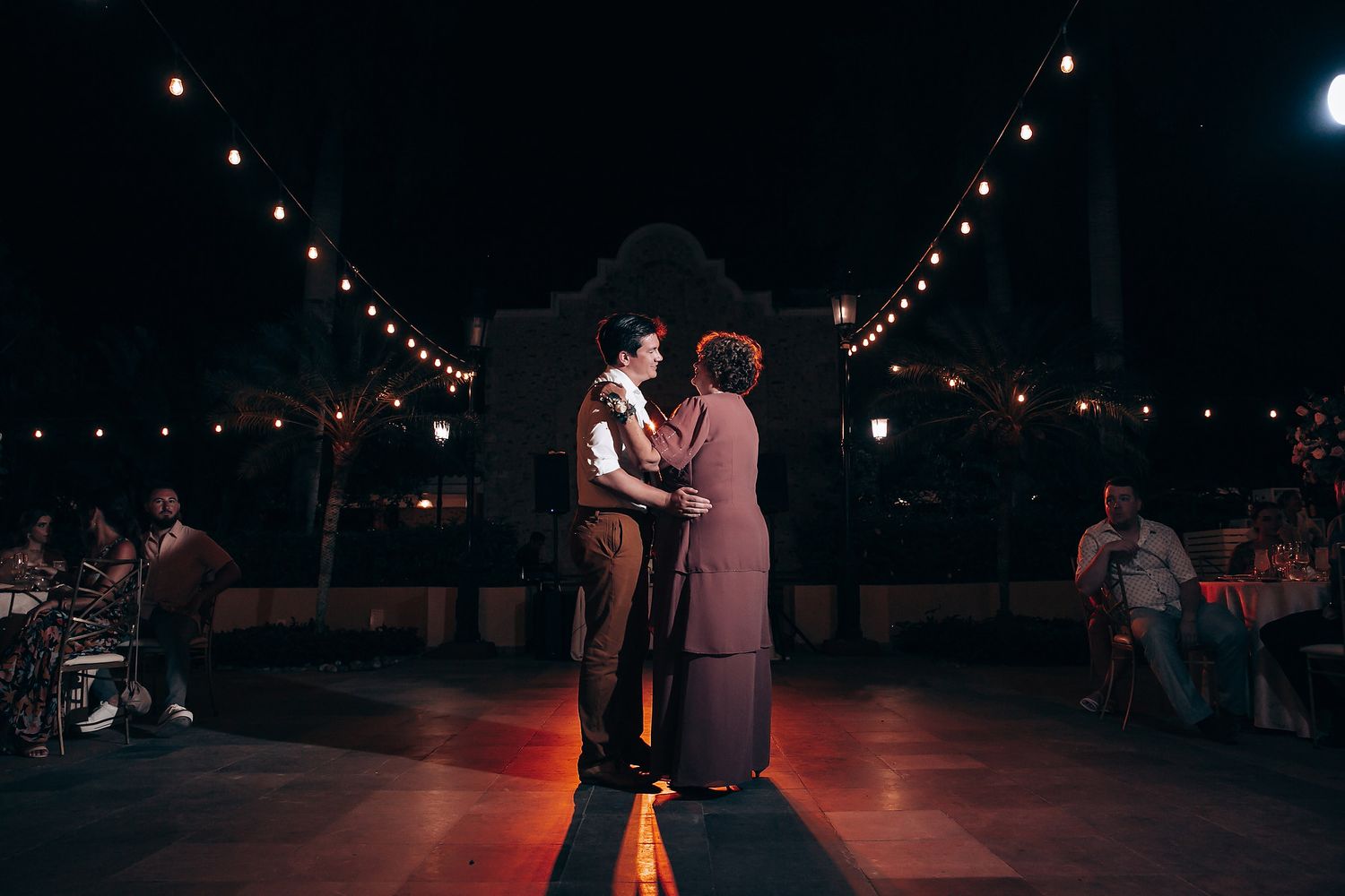 A couple dances together under warm string lights at an outdoor evening reception.