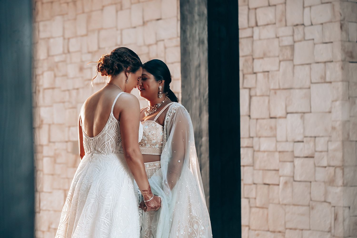 Two people in elegant white wedding dresses share an intimate moment against a stone wall.