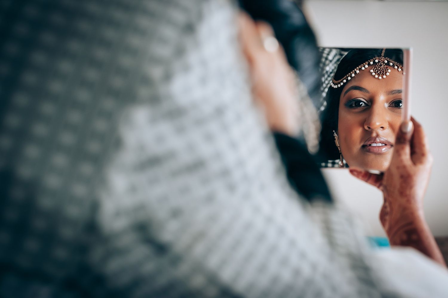 A reflection in a mirror shows a bride wearing a delicate crystal headpiece and elegant bridal jewelry.