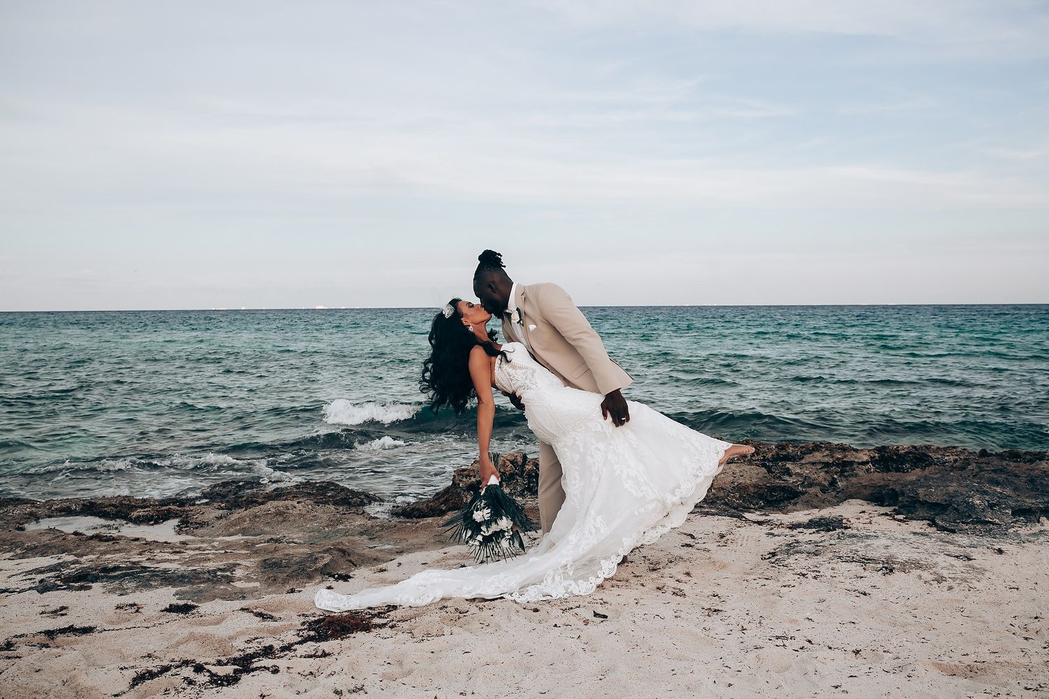 A couple shares a romantic moment on rocky beach shore with flowing white wedding dress.