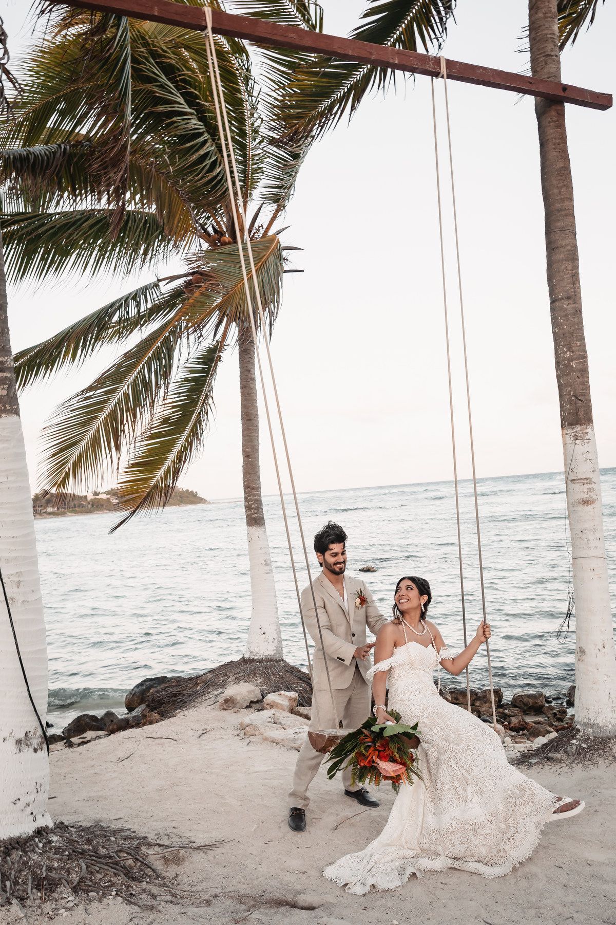 Groom pushing bride on beach swing surrounded by palm trees at sunset.